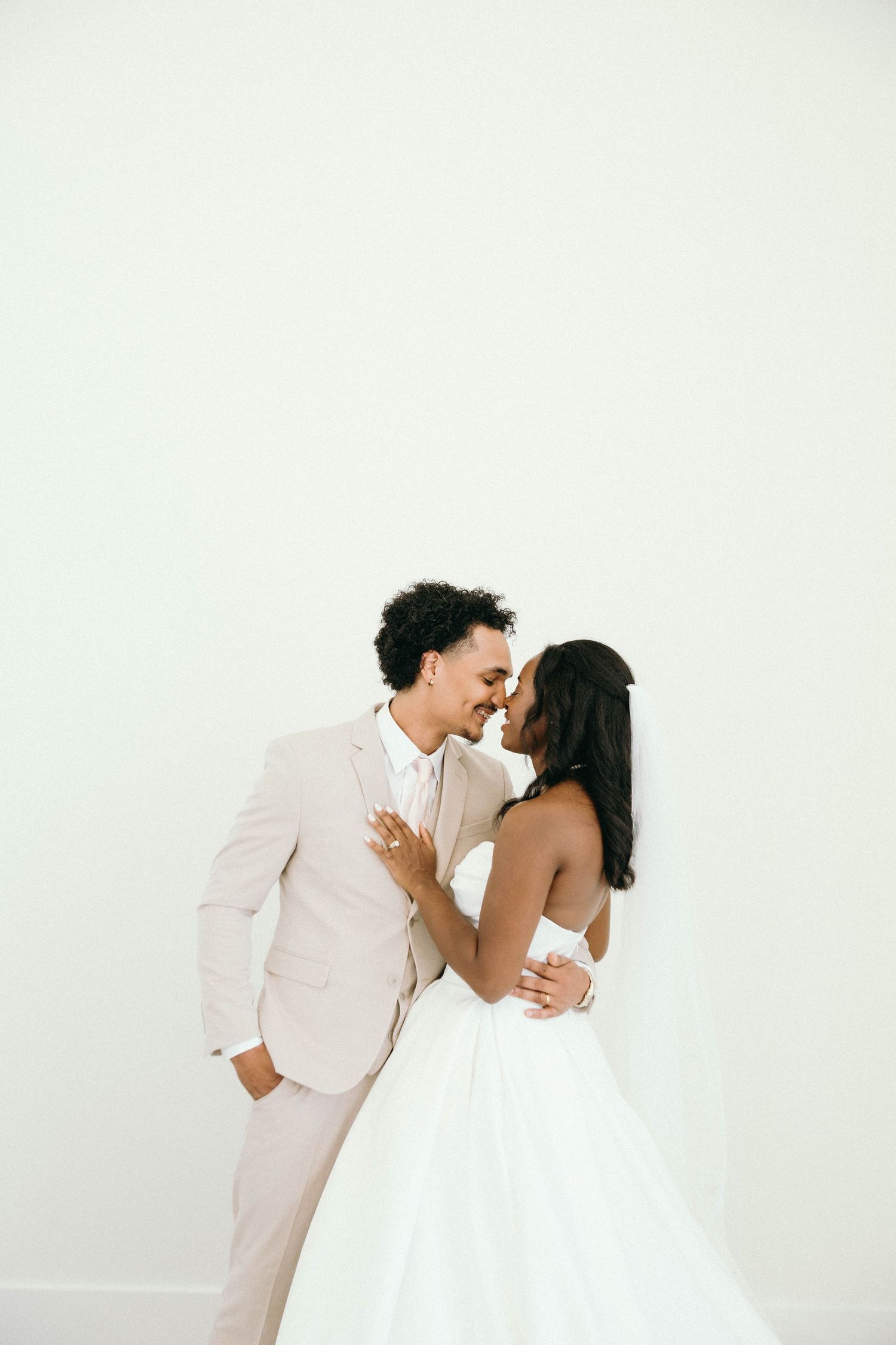 A bride and groom share a romantic moment, touching foreheads, dressed in white gown and blush suit against a white backdrop.