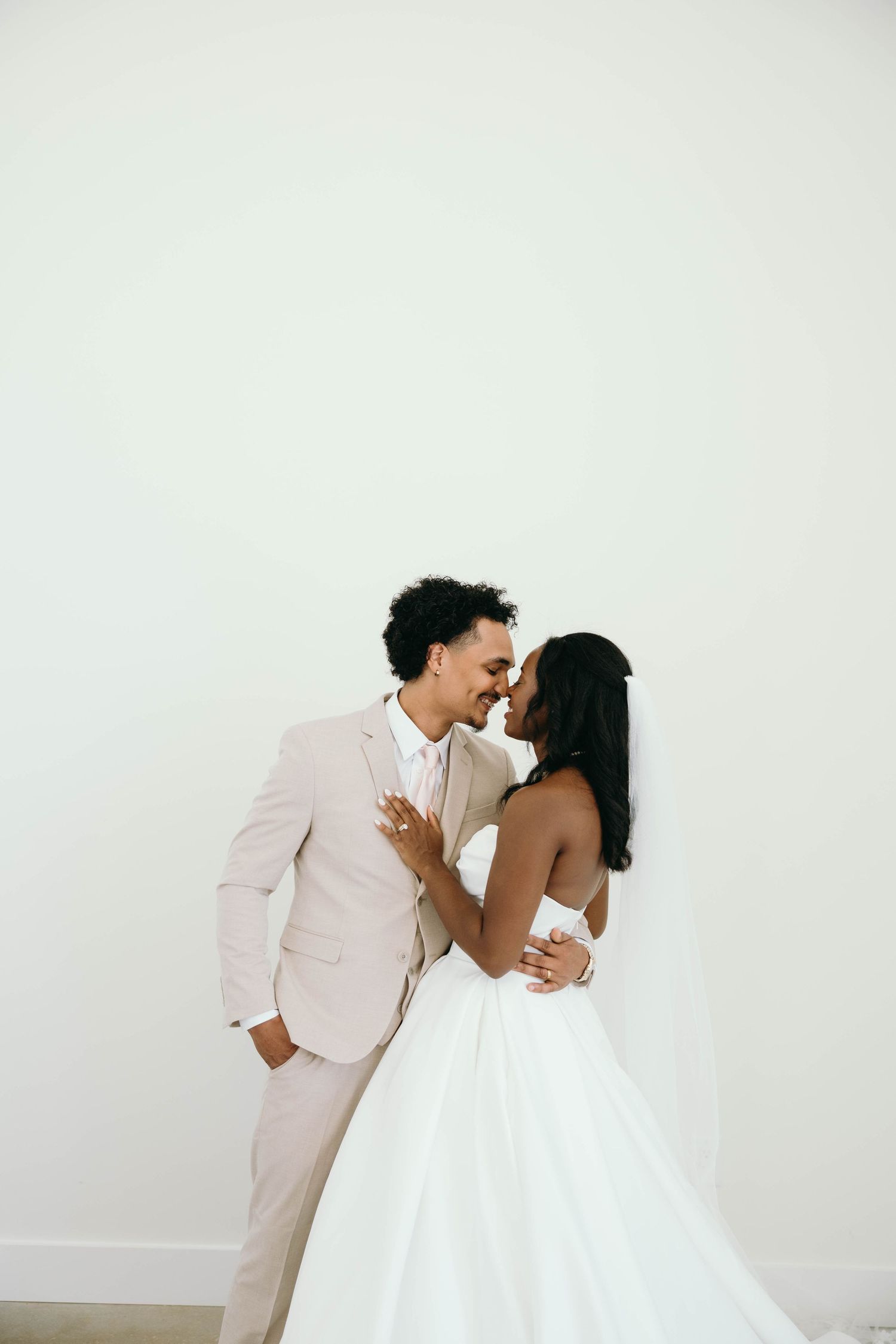 A romantic wedding couple in white attire shares intimate moments in a minimalist setting against a light background.