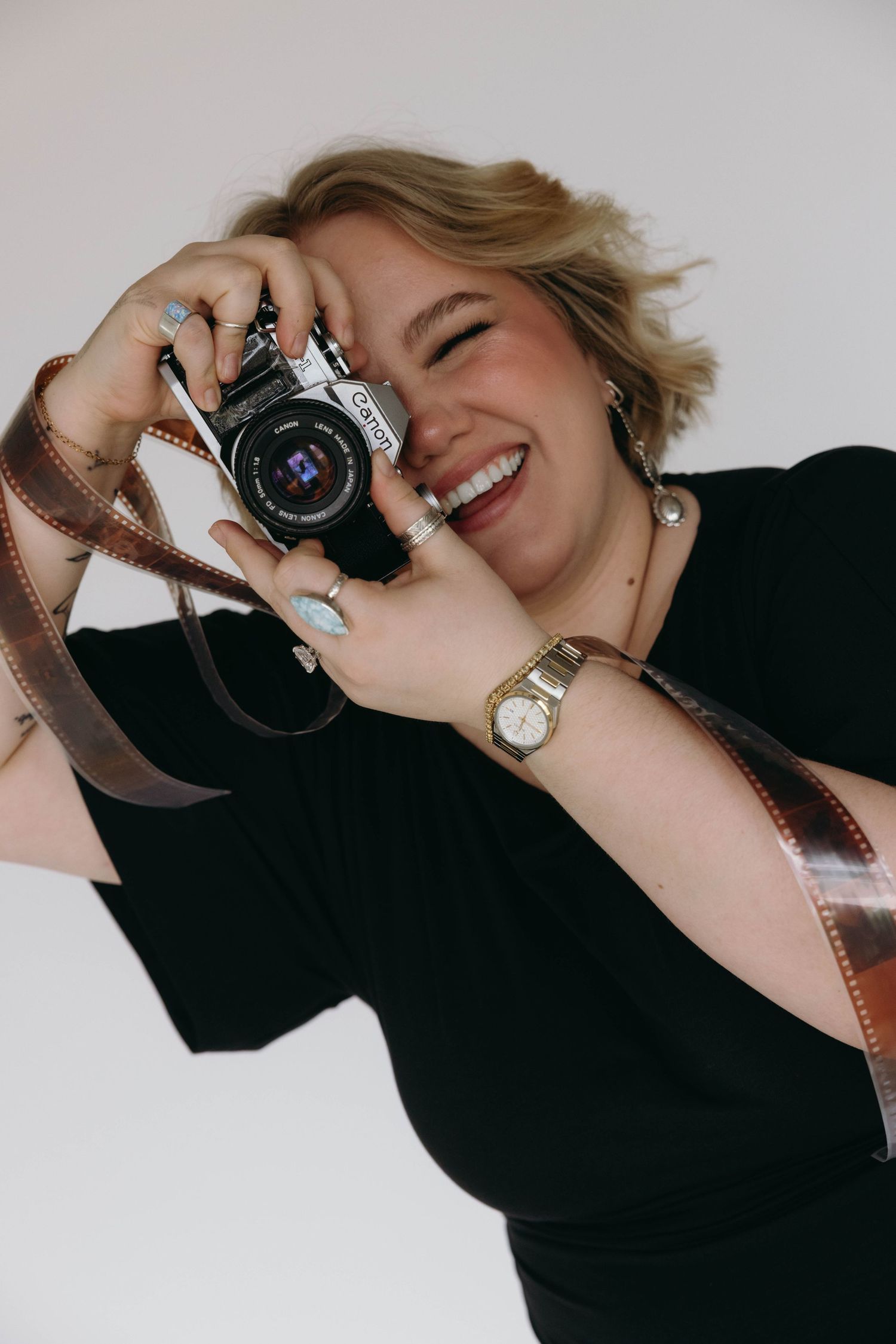 Smiling woman holding a Canon film camera with a film strip draped over her wrist, wearing a gold watch and rings.