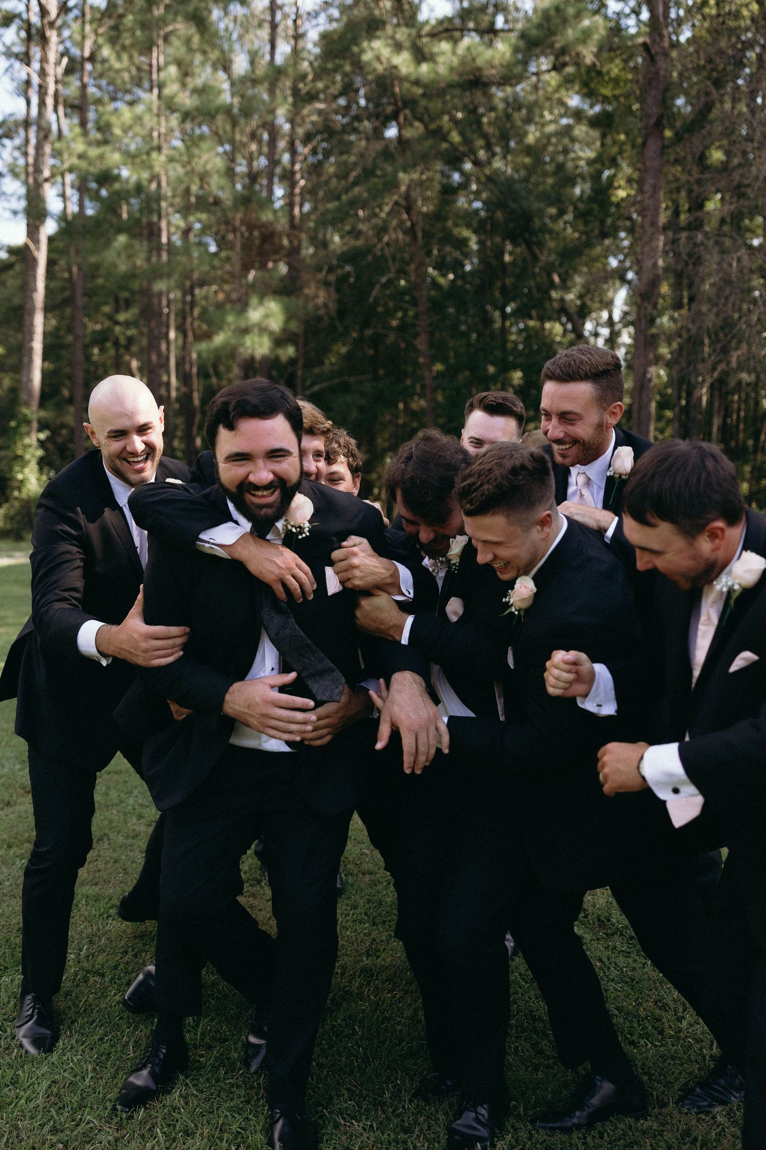 A group of groomsmen in black suits playfully horsing around and laughing together on a grassy lawn near pine trees.