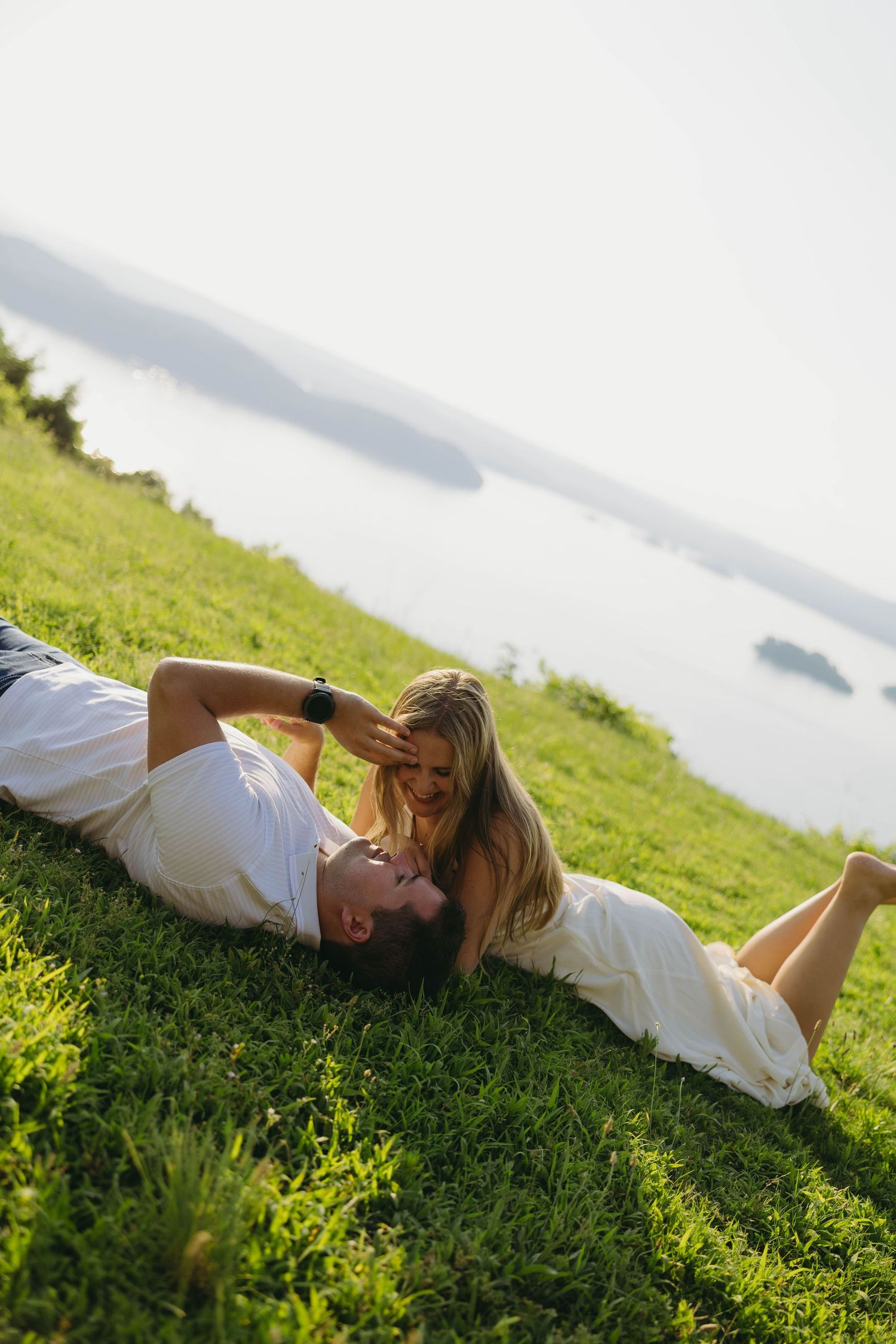 Couple lying on green grass hillside with scenic lake view, man looking up at smiling blonde woman in white dress.