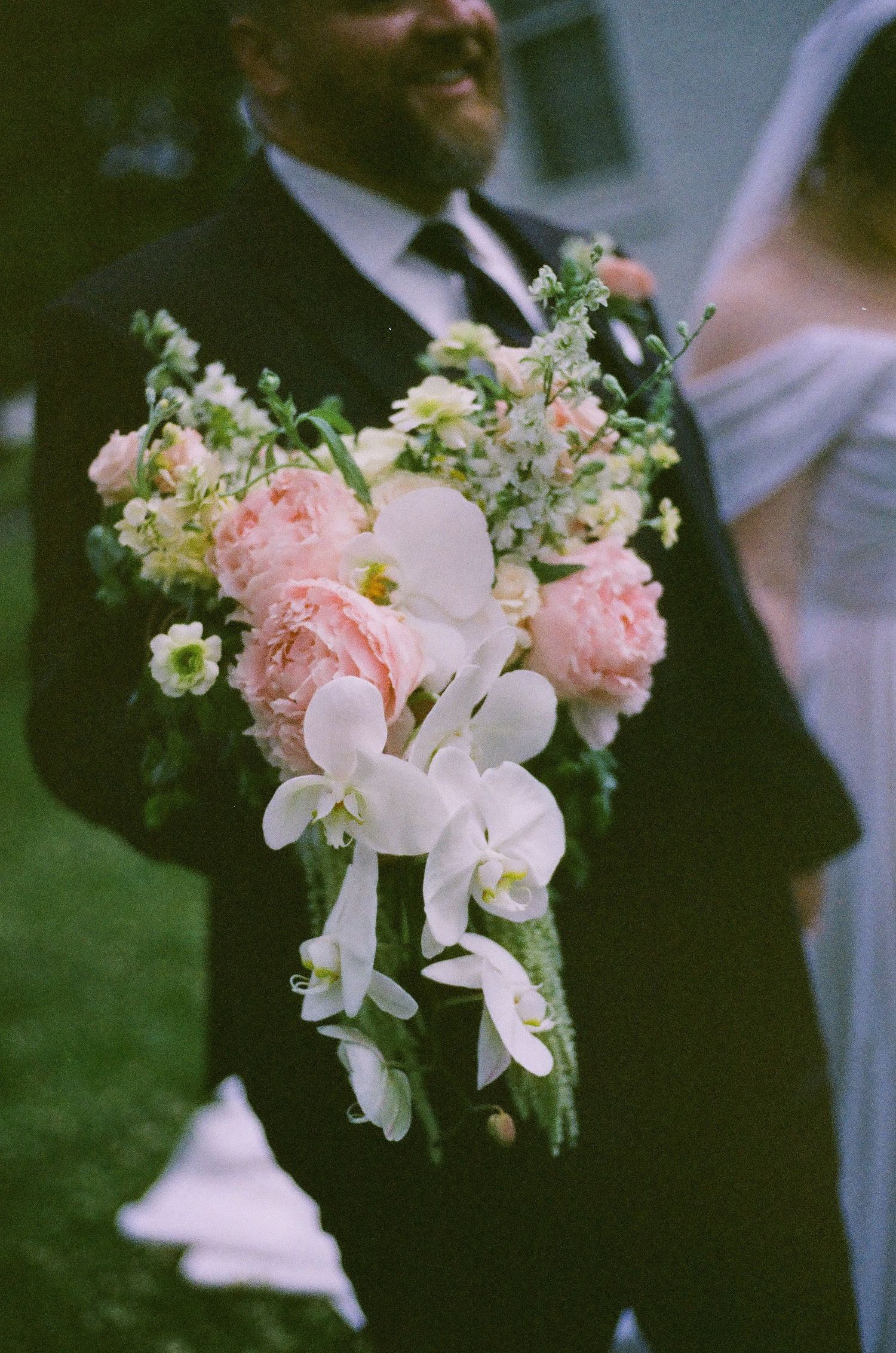 A groom in a black suit holds a bridal bouquet of pink peonies, white orchids, and greenery at a wedding.