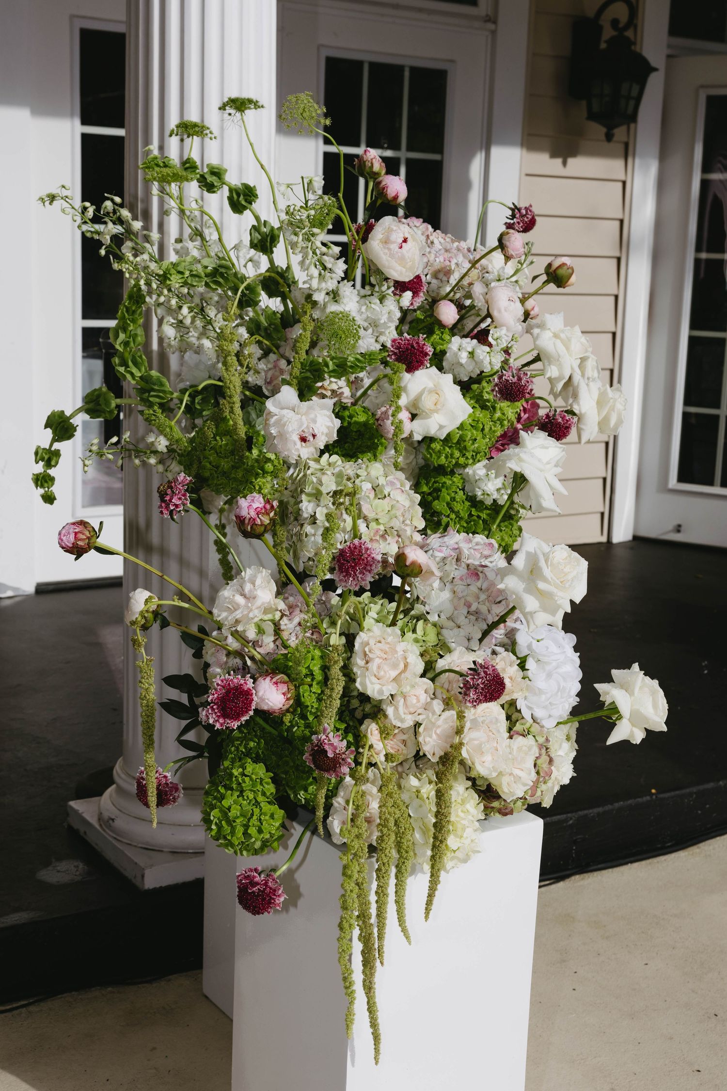 Elegant white and burgundy floral arrangements in tall white vases line a covered porch with white columns.