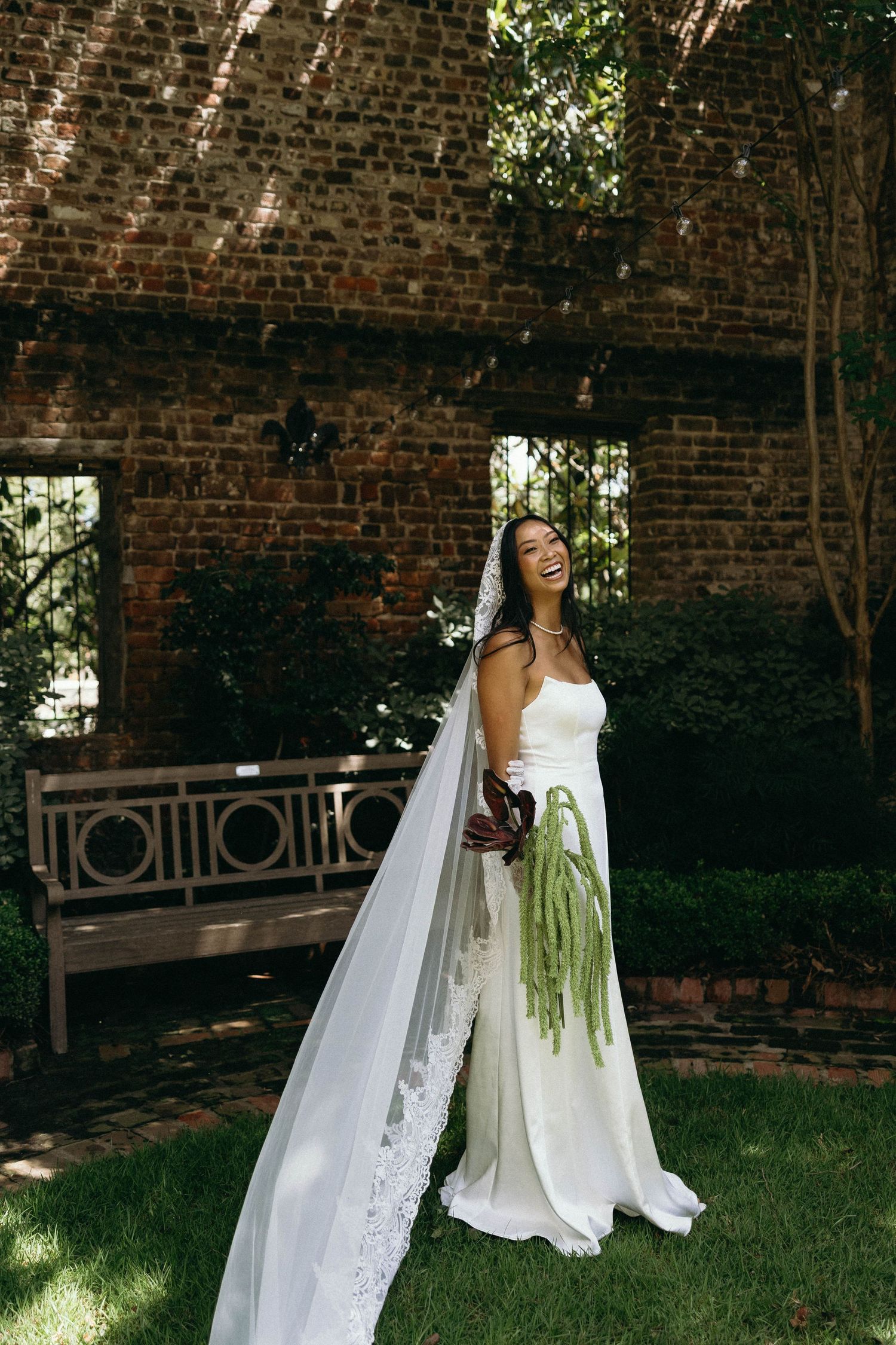 A bride in a white wedding dress holds a cascading green bouquet while standing near a brick wall in an outdoor garden setting.