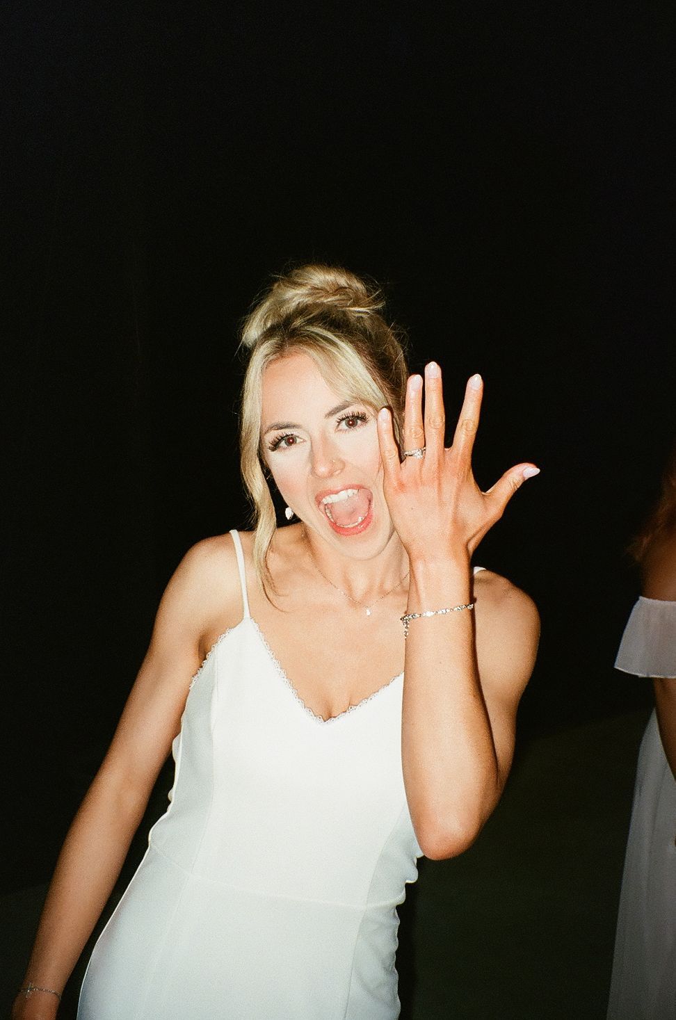 Excited bride in white dress shows off engagement ring while making joyful expression.