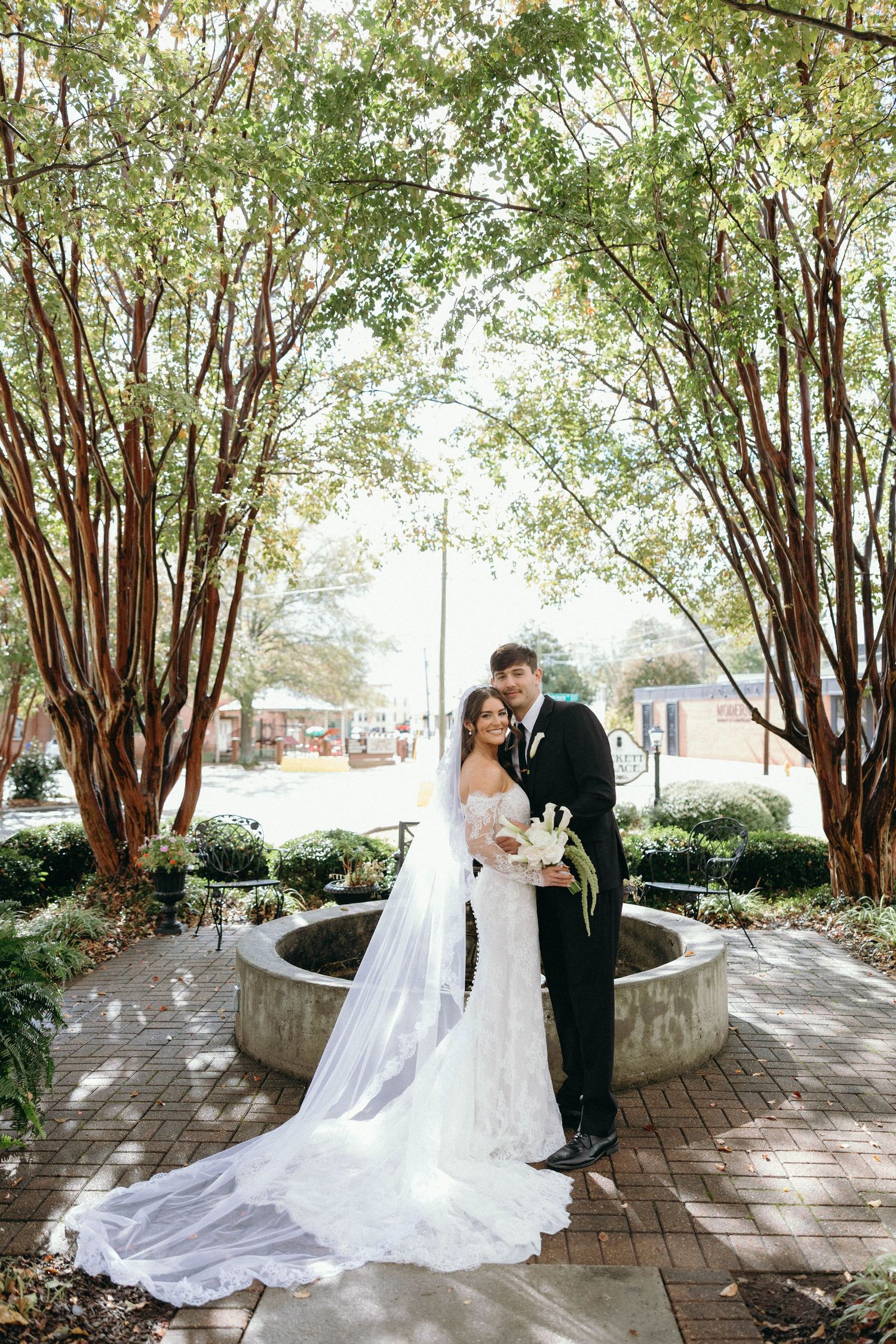 Bride and groom pose by a stone fountain surrounded by lush green trees on their wedding day.