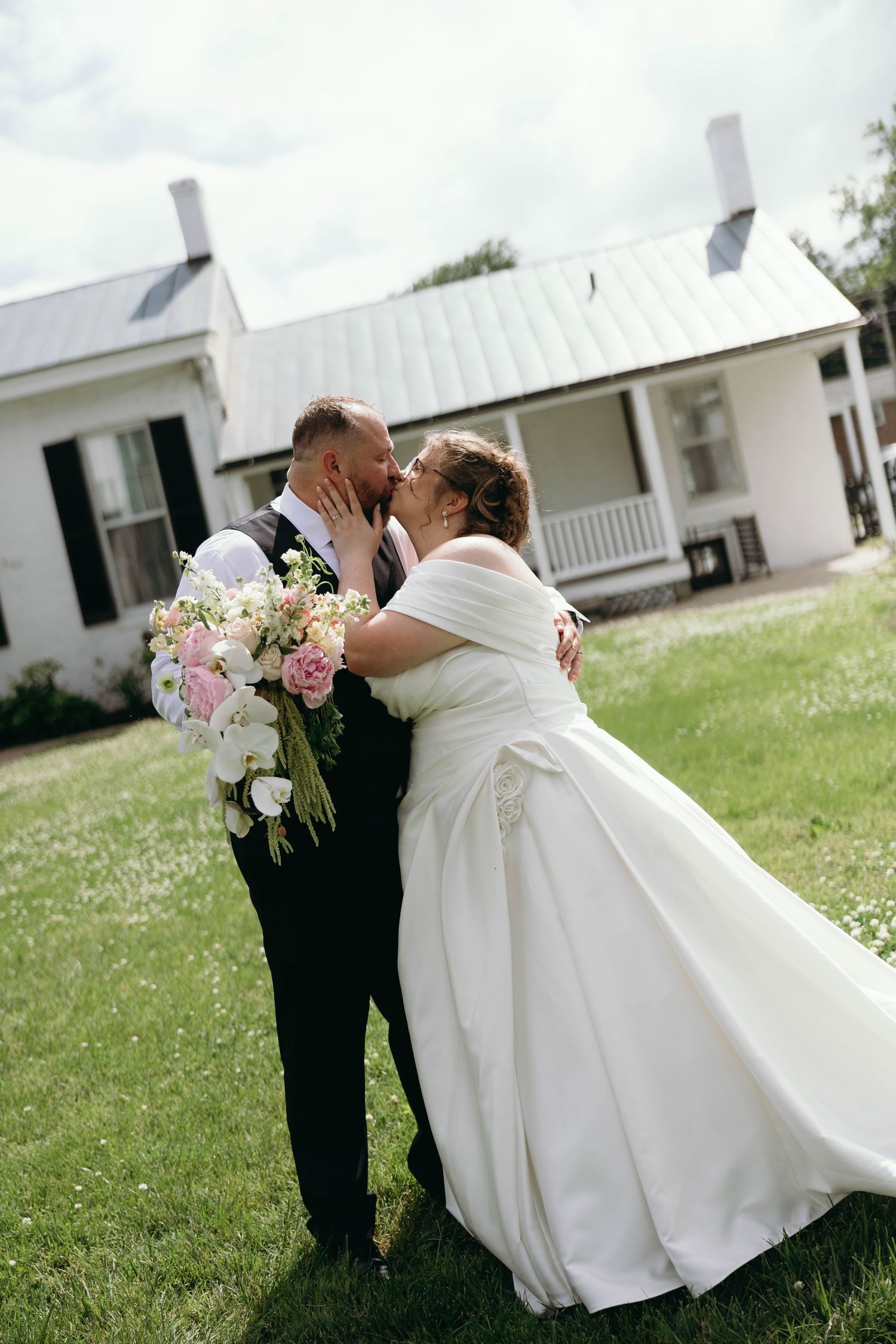 Bride and groom share a kiss on green lawn outside white farmhouse, bride holding pink and white bouquet in wedding dress.