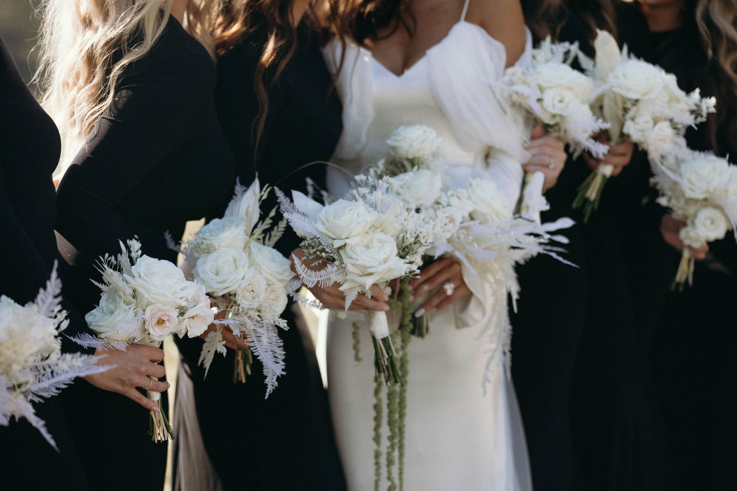 Wedding party members in black dresses hold elegant white and cream floral bouquets with feather accents.