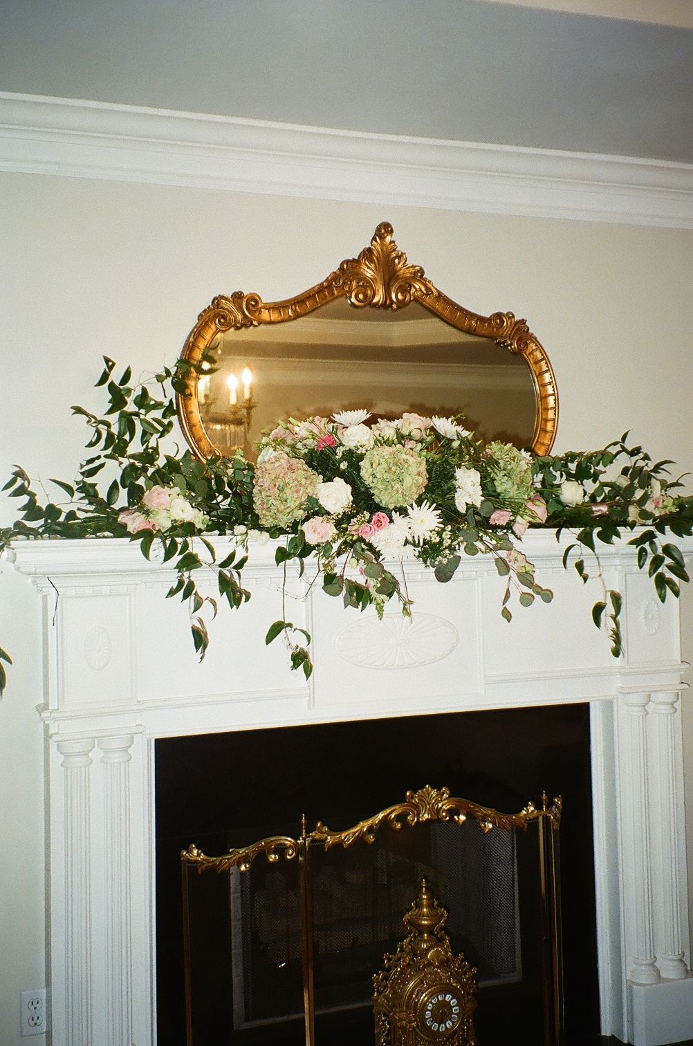 Ornate gold mirror hangs above white fireplace mantle decorated with lush greenery and white hydrangea floral arrangement.