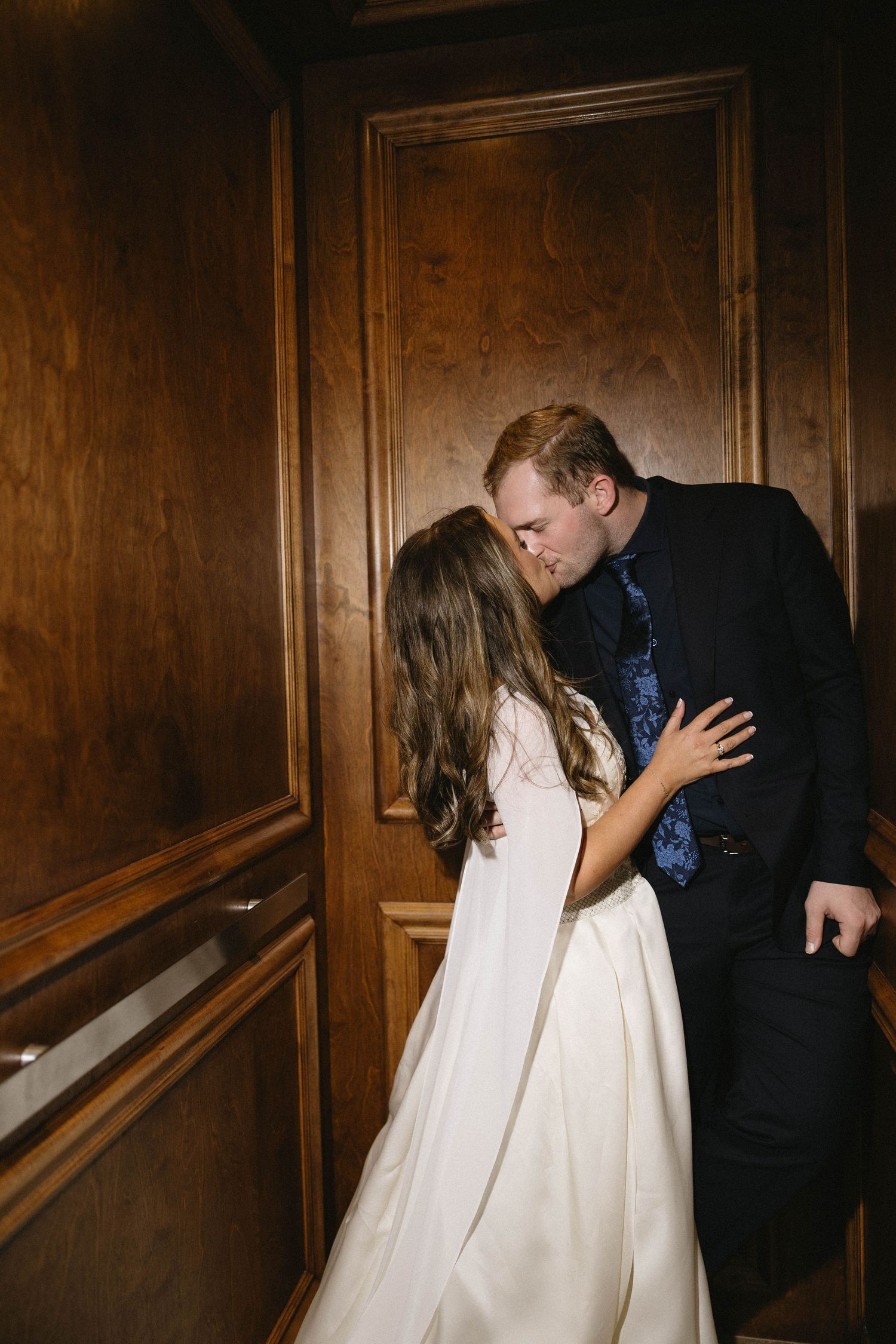Bride and groom sharing a romantic kiss in an elegant wood-paneled hallway during their wedding reception.