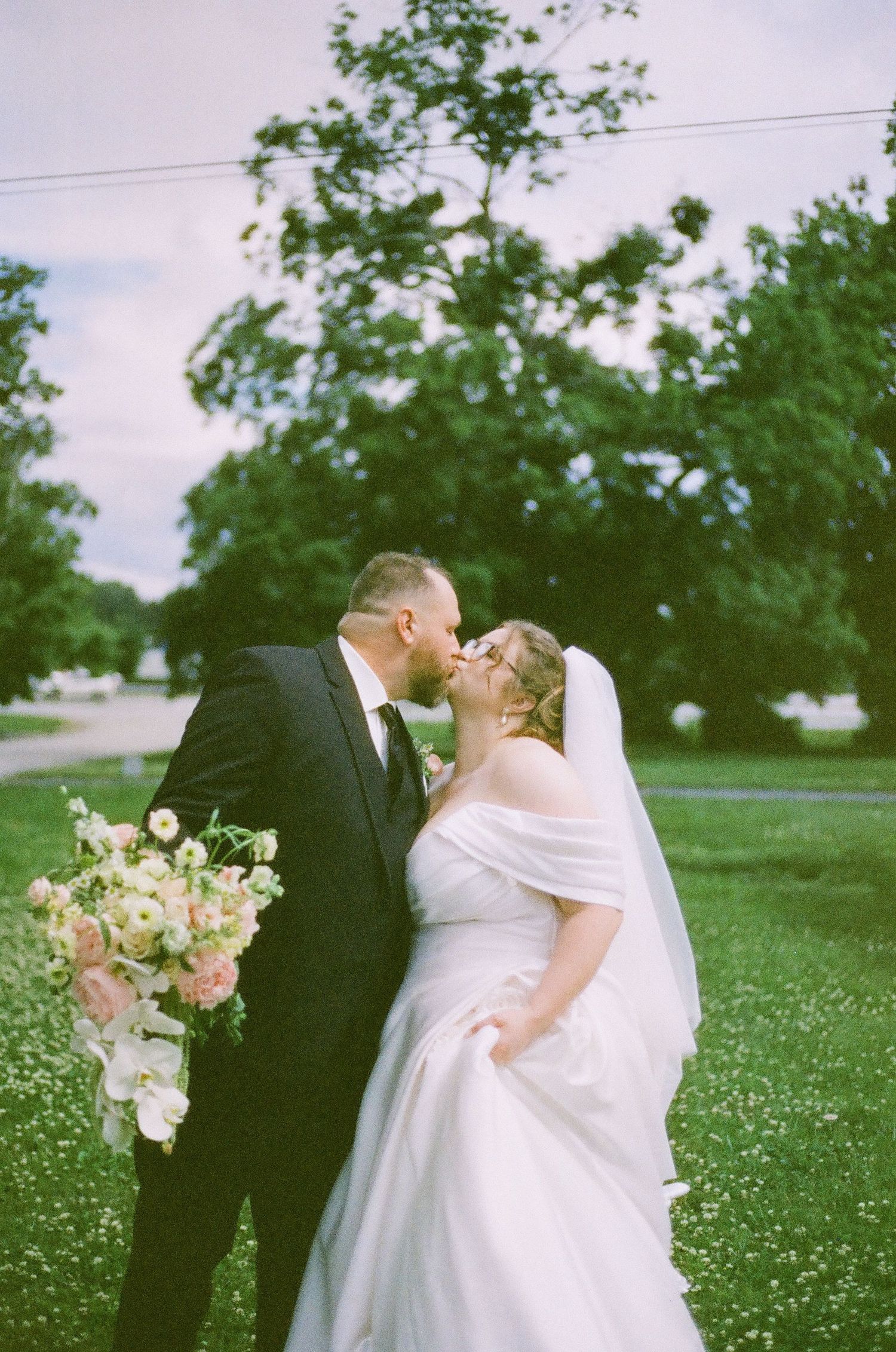 A bride and groom share a romantic kiss outdoors on their wedding day, surrounded by lush green trees.