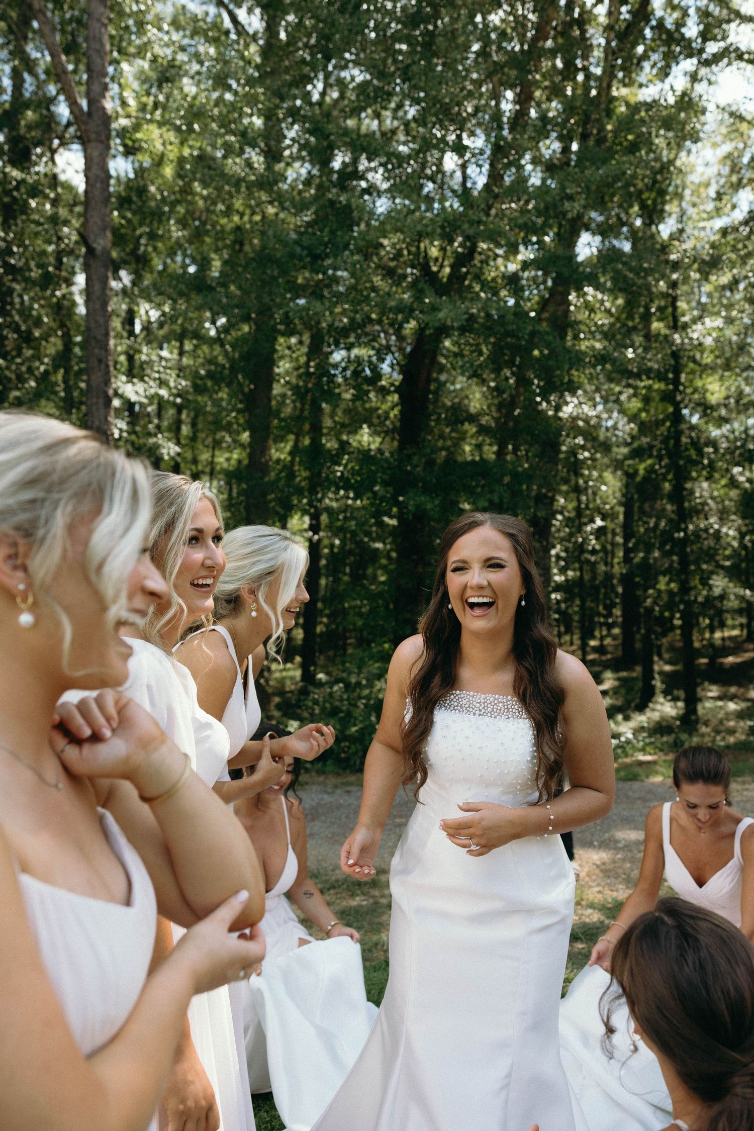 A joyful outdoor wedding scene with bridesmaids in white dresses laughing together in a wooded setting.