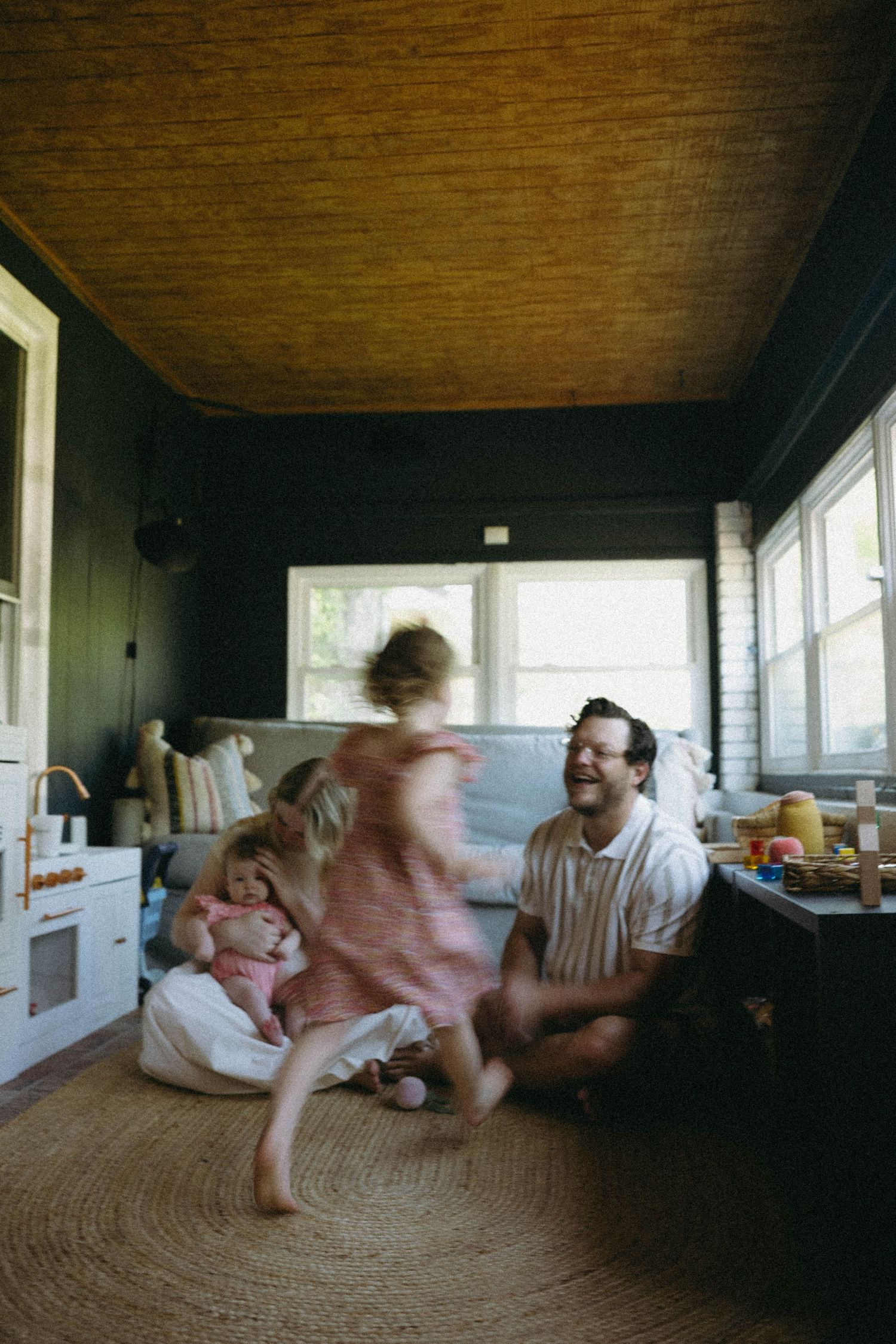 A father smiles as his young daughter twirls on a jute rug while another child sits nearby in a cozy, dark-walled room.