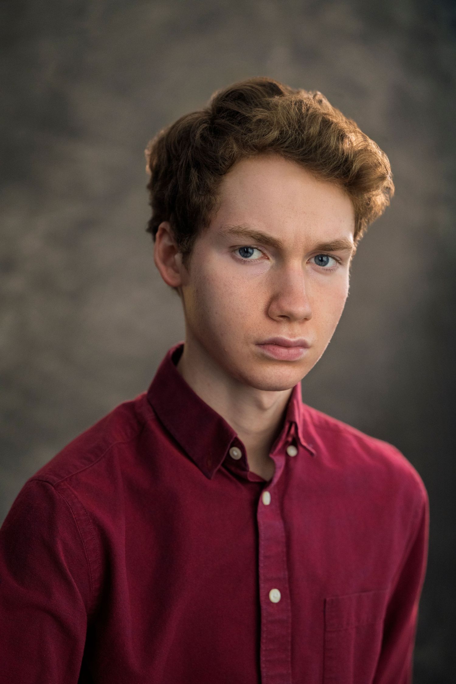 Young man with red curly hair and blue eyes wearing a dark red button-up shirt posed for a portrait photo.