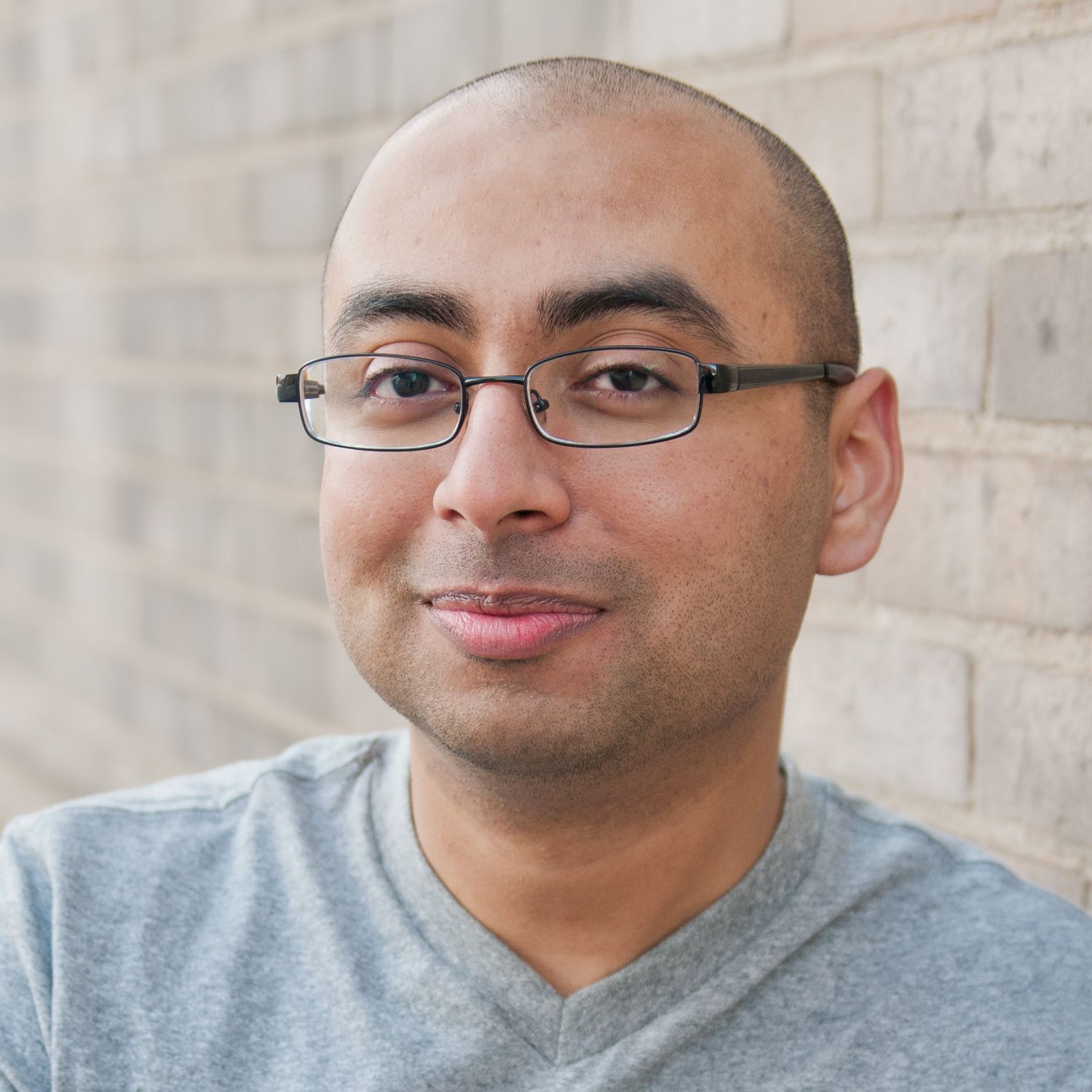 Professional headshot of a smiling bald man wearing glasses and a gray t-shirt against a brick wall background.