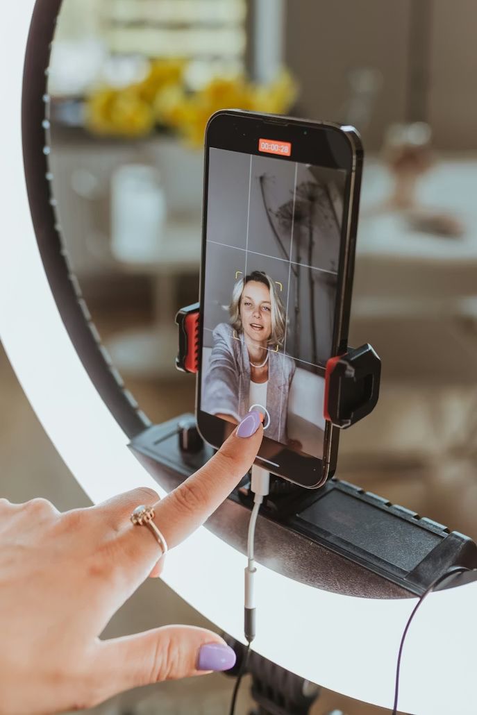 Woman recording herself on a smartphone mounted on a ring light stand for social media content creation.
