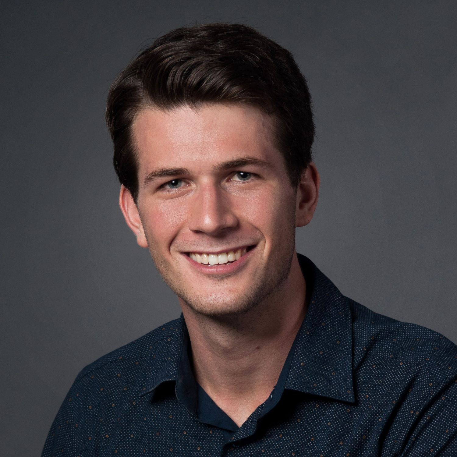 Professional headshot of a young smiling man with dark hair wearing a dark blue polo shirt against a gray background.