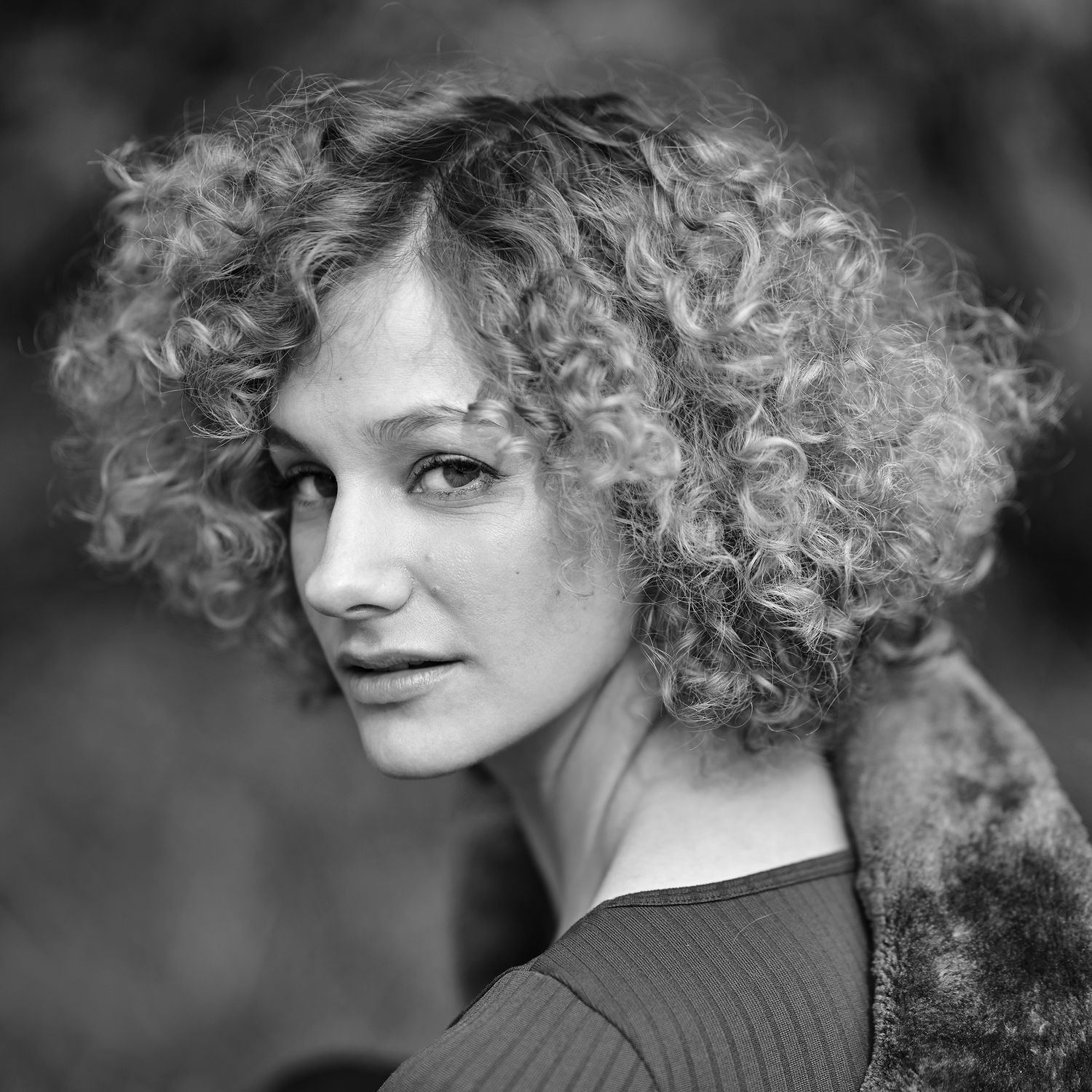 Black and white portrait of a young woman with curly hair looking over her shoulder outdoors.