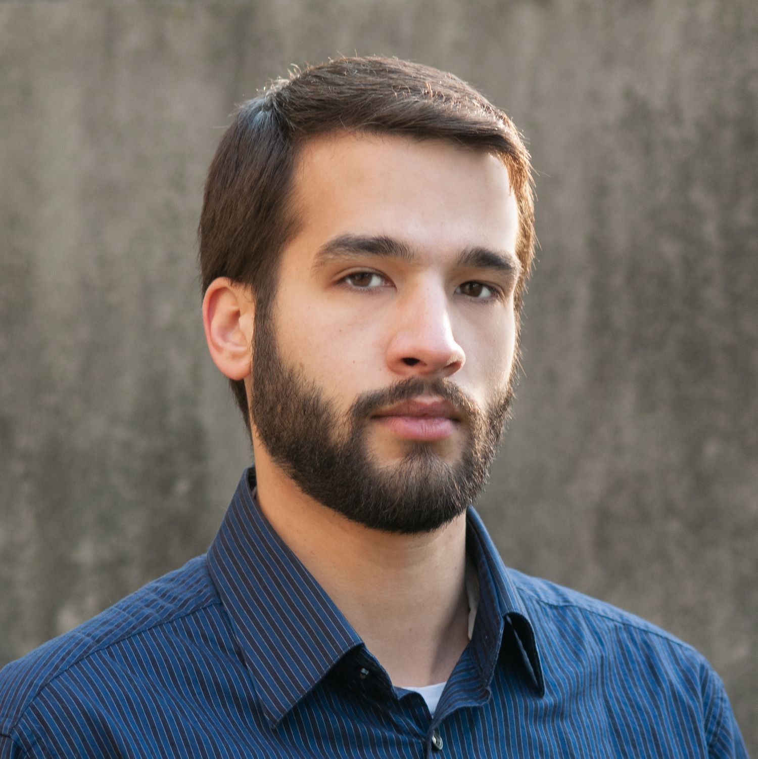 Professional headshot of a young bearded man with dark hair wearing a blue striped shirt against a concrete background.