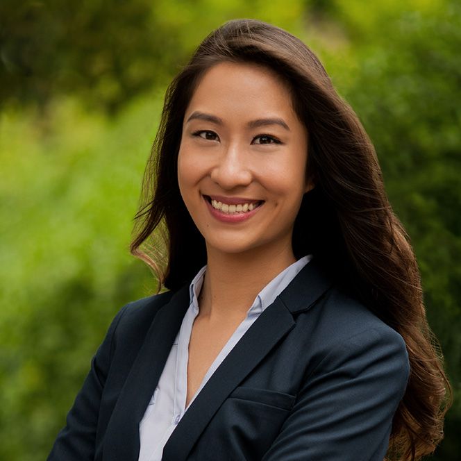 Smiling young Asian woman in a navy blazer poses outdoors with green foliage background.