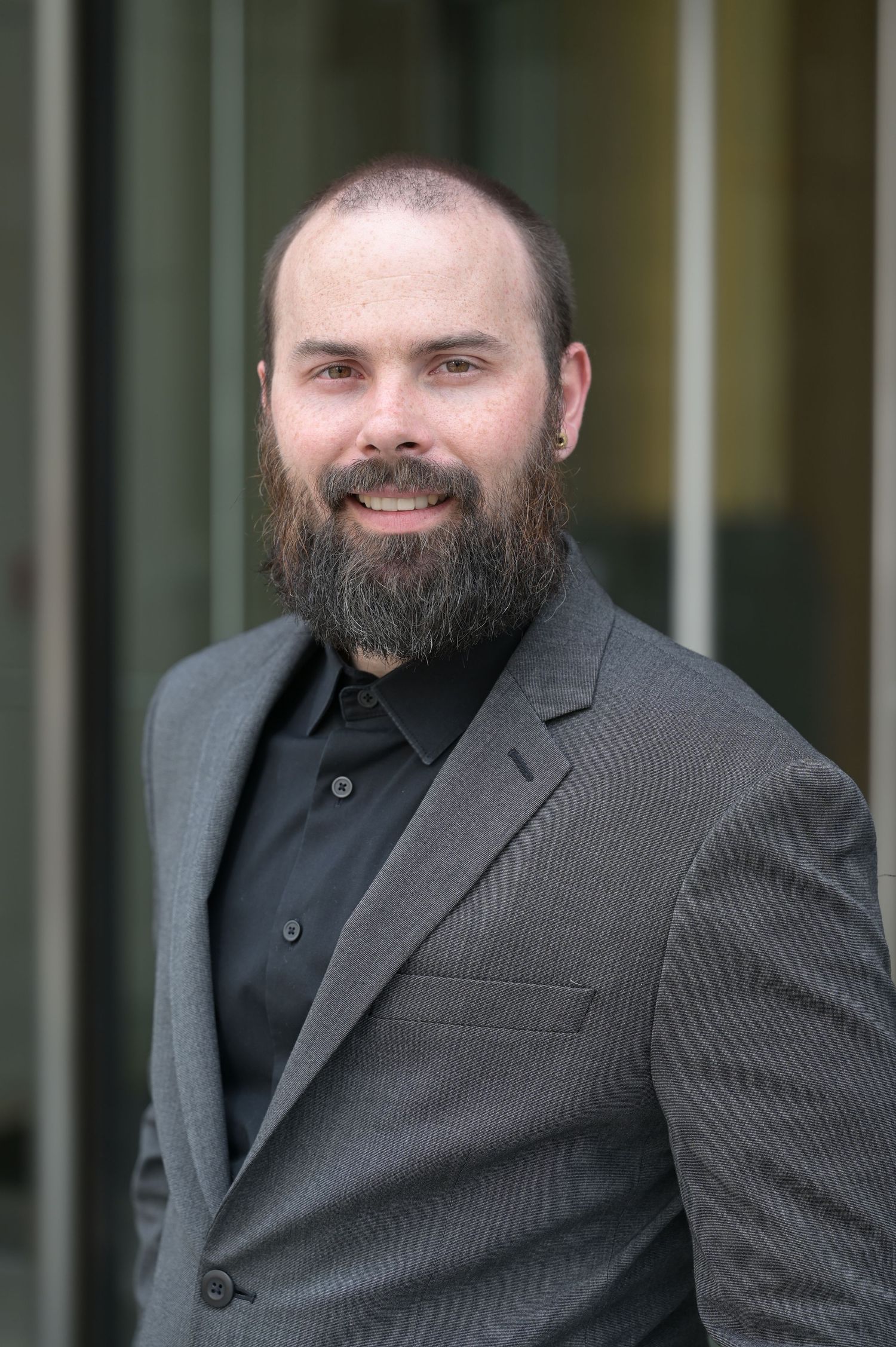 Professional man with a beard wearing a gray suit jacket posing outdoors.