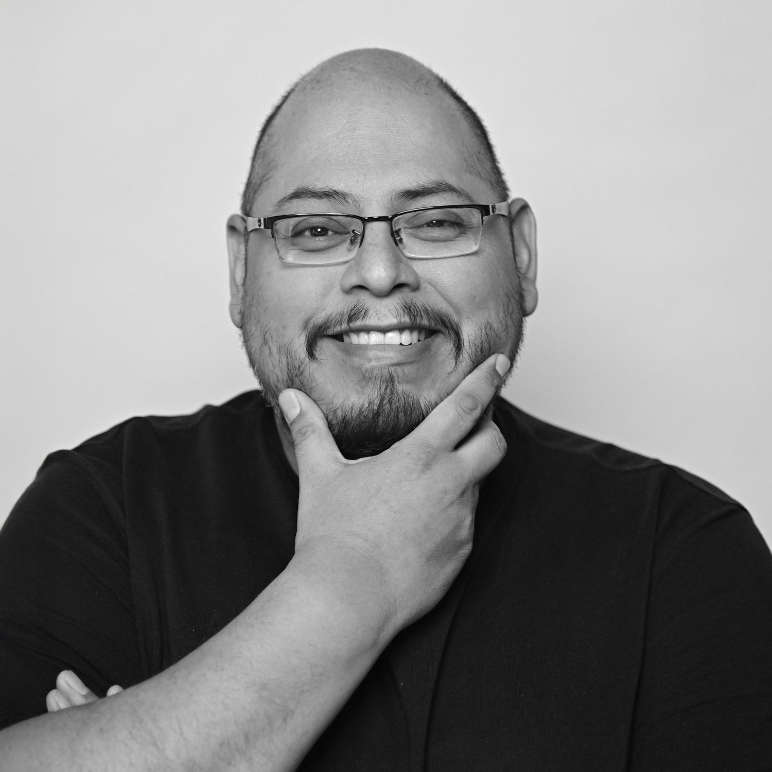 Black and white portrait of a smiling bald man with glasses and a goatee, posing thoughtfully with hand on chin.