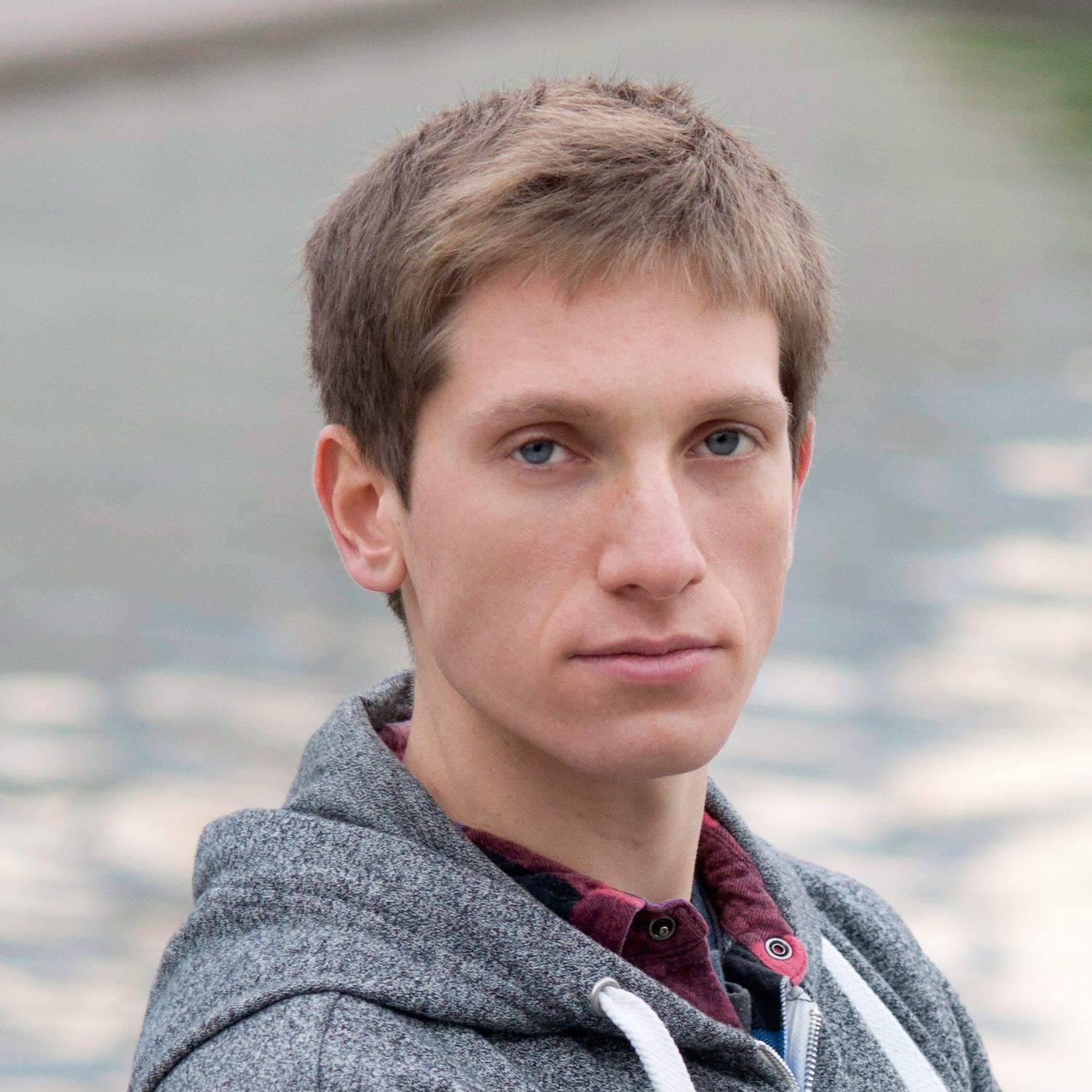 Professional headshot of a young man with short blonde hair wearing a gray hoodie outdoors near water.