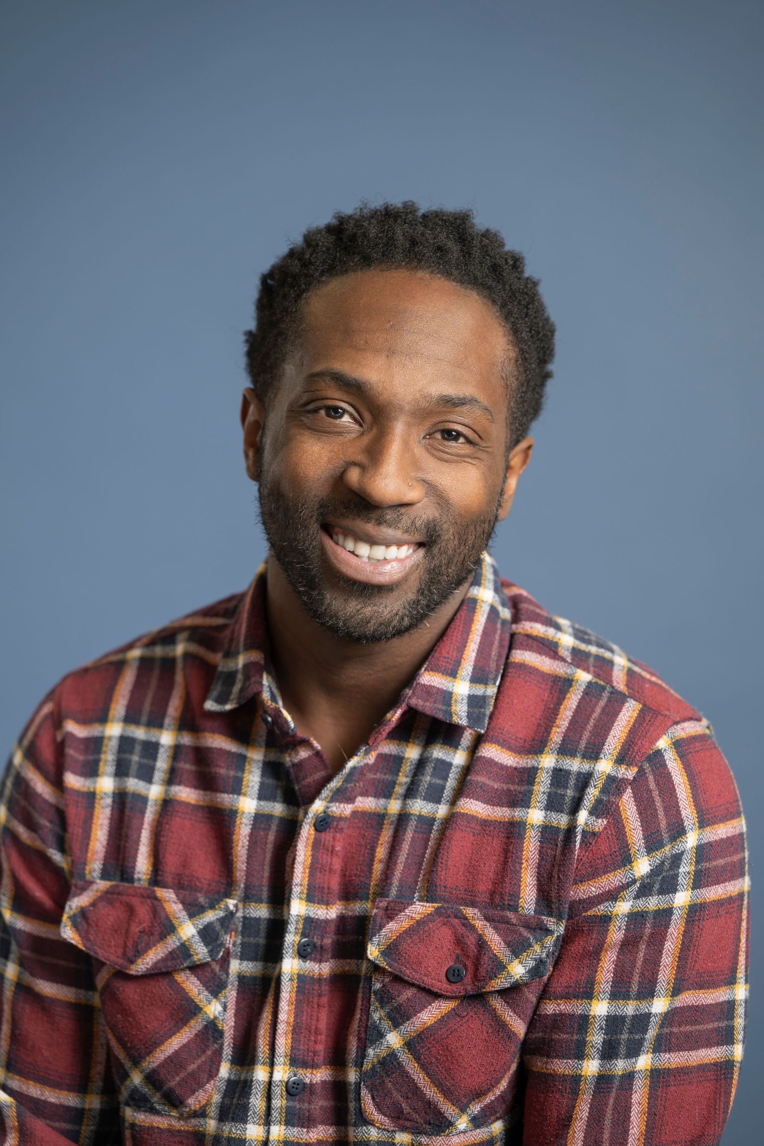 Smiling Black man with short hair and beard wearing a red plaid flannel shirt against a blue background.