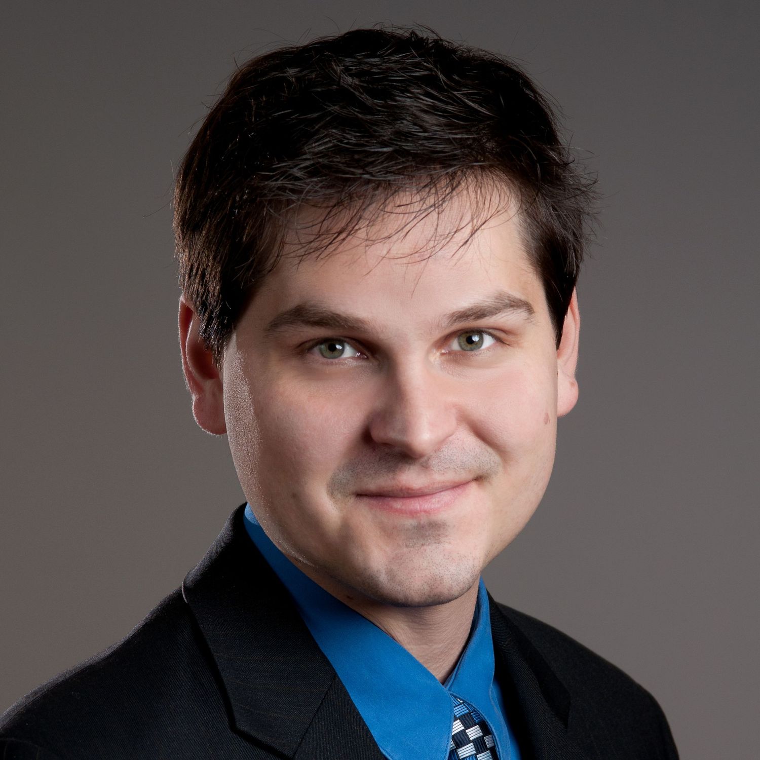Professional headshot of a smiling young man with dark hair wearing a blue shirt and black blazer on gray background.