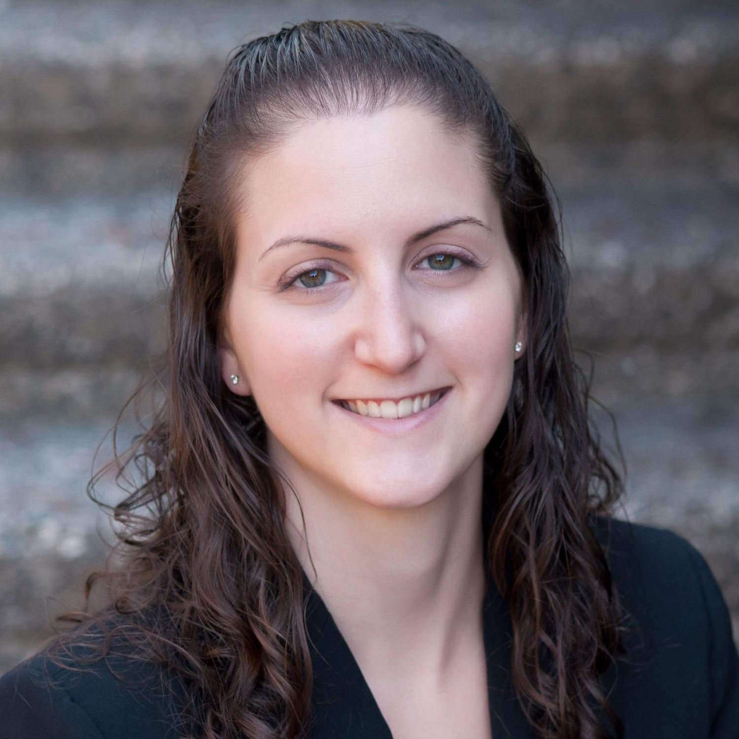 Professional headshot of a smiling young woman with long curly dark hair wearing a black blazer on stone steps.