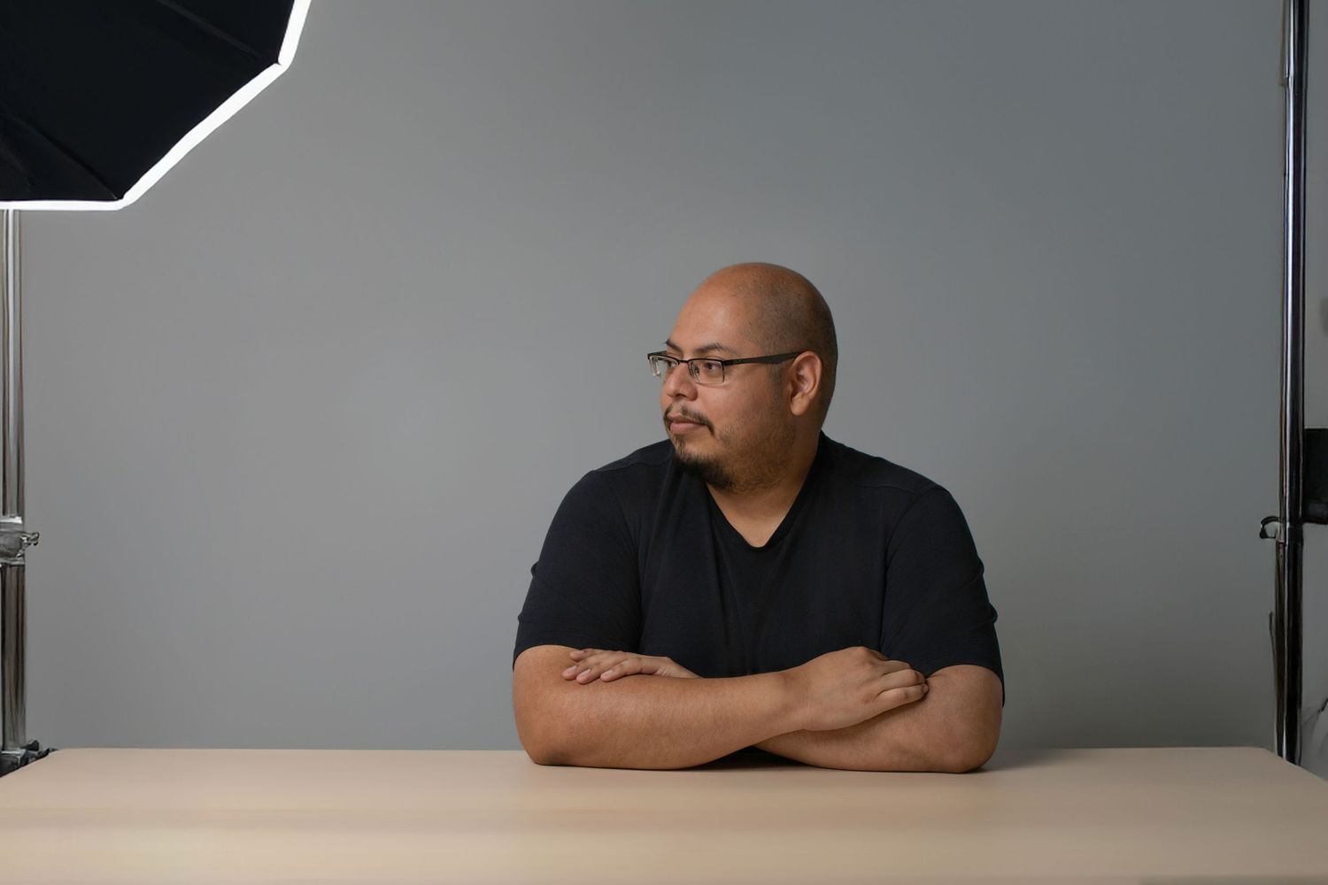 Bald man with glasses sits with arms crossed at a table in a photography studio.