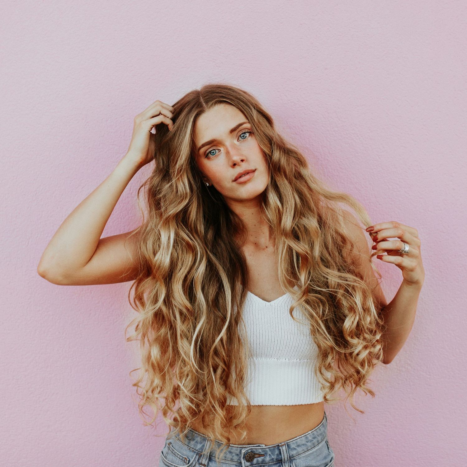 Young woman with long wavy blonde hair poses against a pink wall wearing a white crop top and jeans.