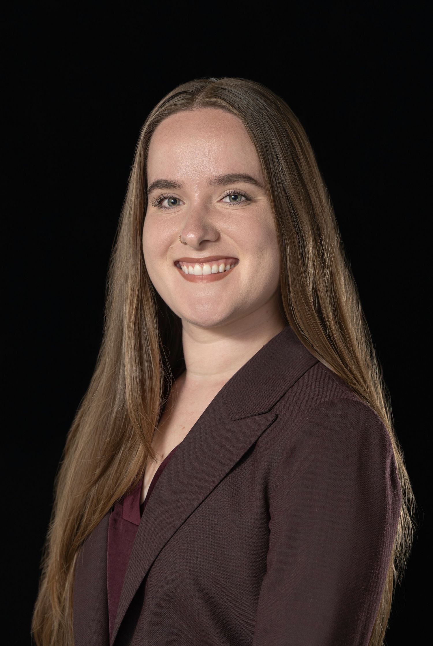 Young professional woman with long brown hair smiling, wearing a dark maroon blazer against a black background.