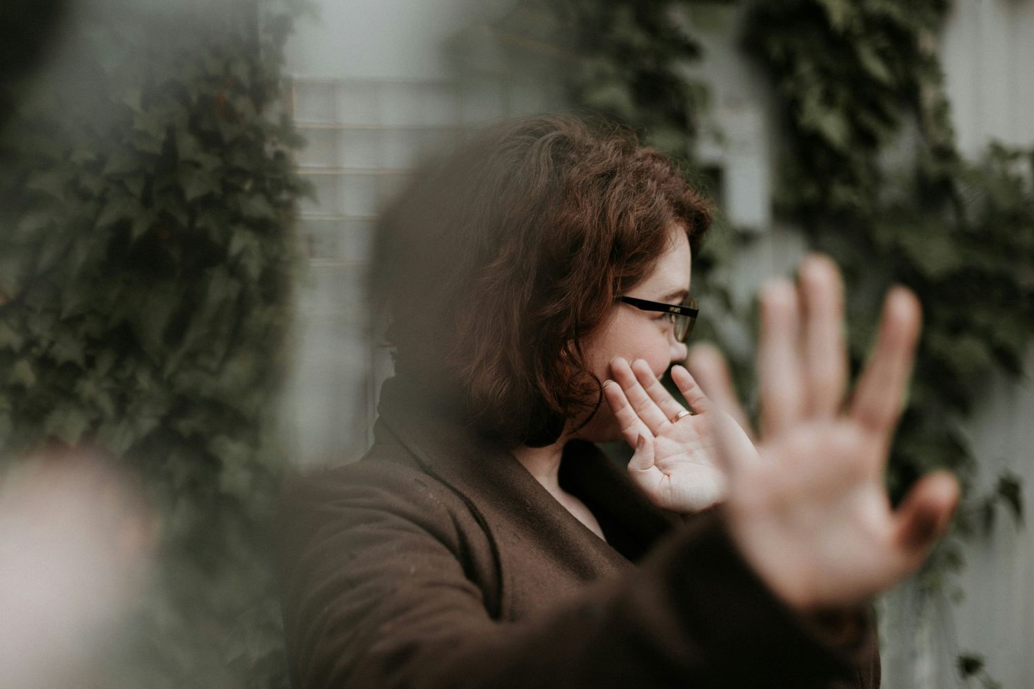 Woman with glasses covering her face with hands outdoors, appearing distressed near ivy-covered wall.