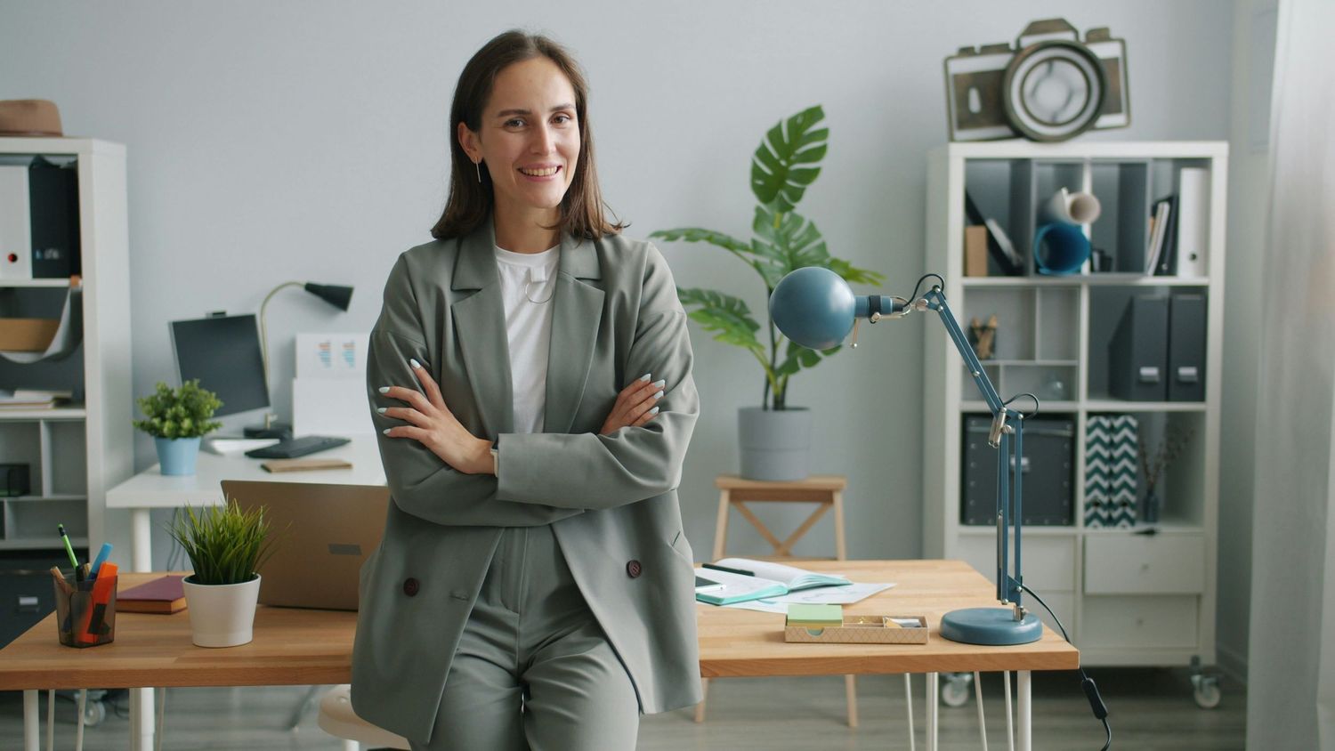 Confident businesswoman in gray suit standing with arms crossed in modern office workspace.