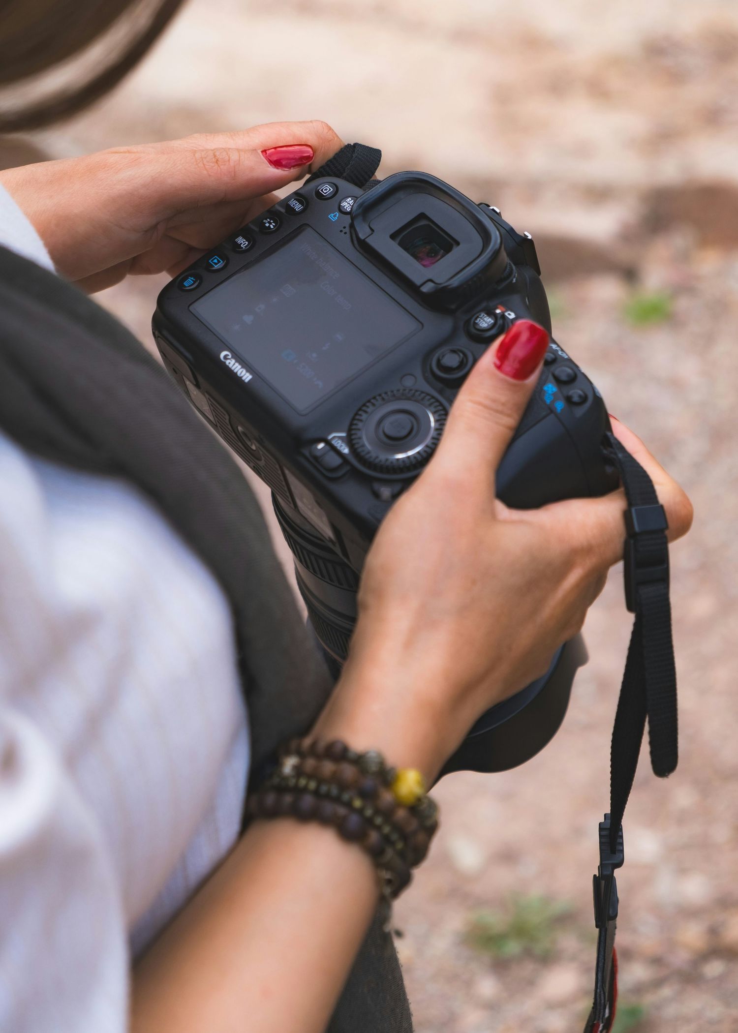 Woman with red nails holding a Canon DSLR camera, wearing a beaded bracelet, reviewing photos outdoors.