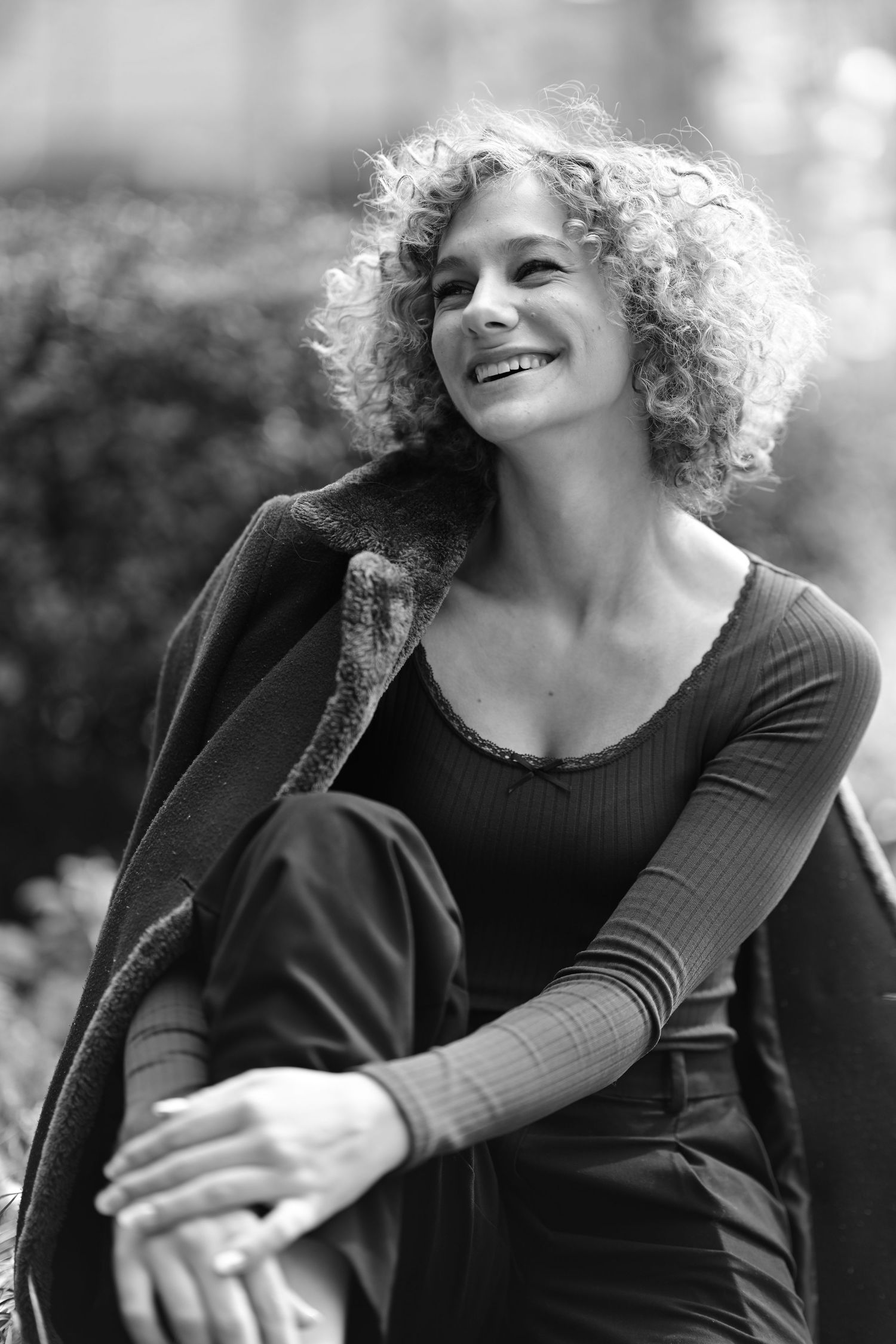 Black and white portrait of a smiling curly-haired woman sitting outdoors in a fitted top and coat.