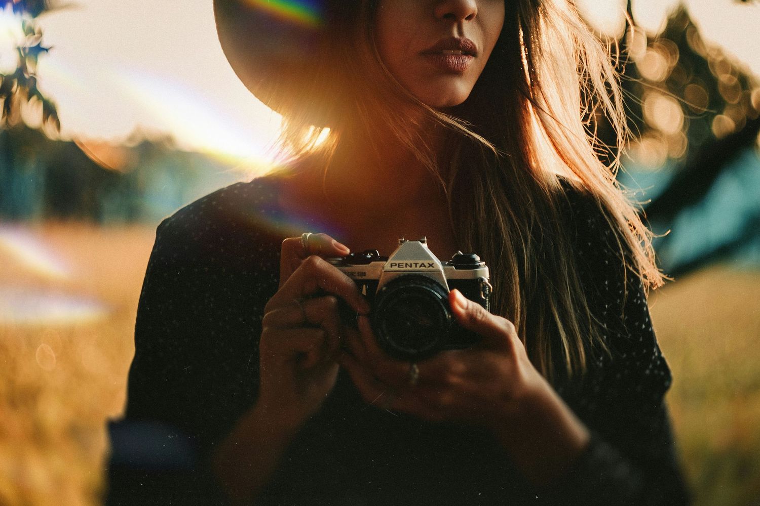 Young woman in wide-brim hat holding a vintage film camera outdoors in golden sunset light.