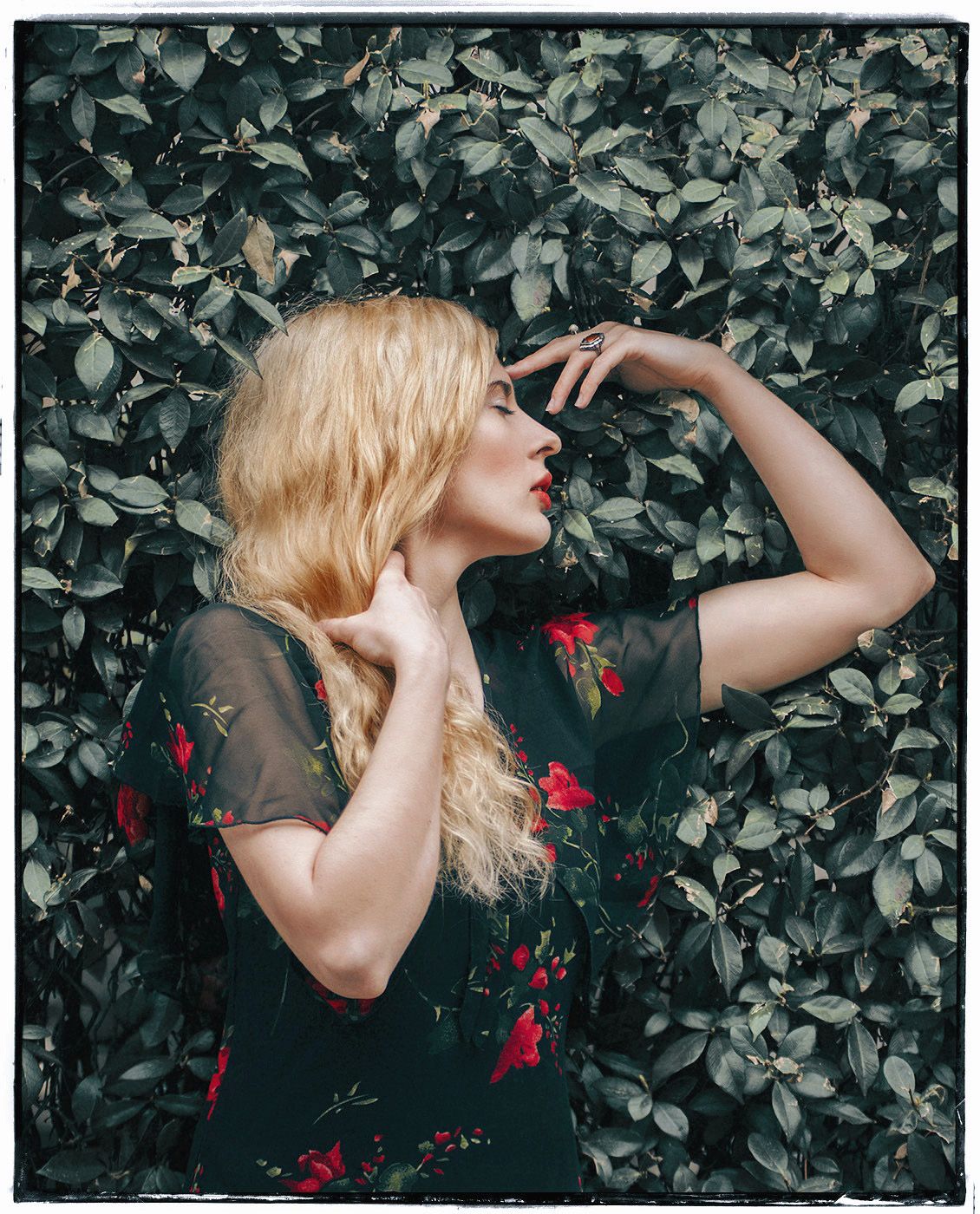 Blonde woman in black floral dress posing against lush green leaf wall outdoors.