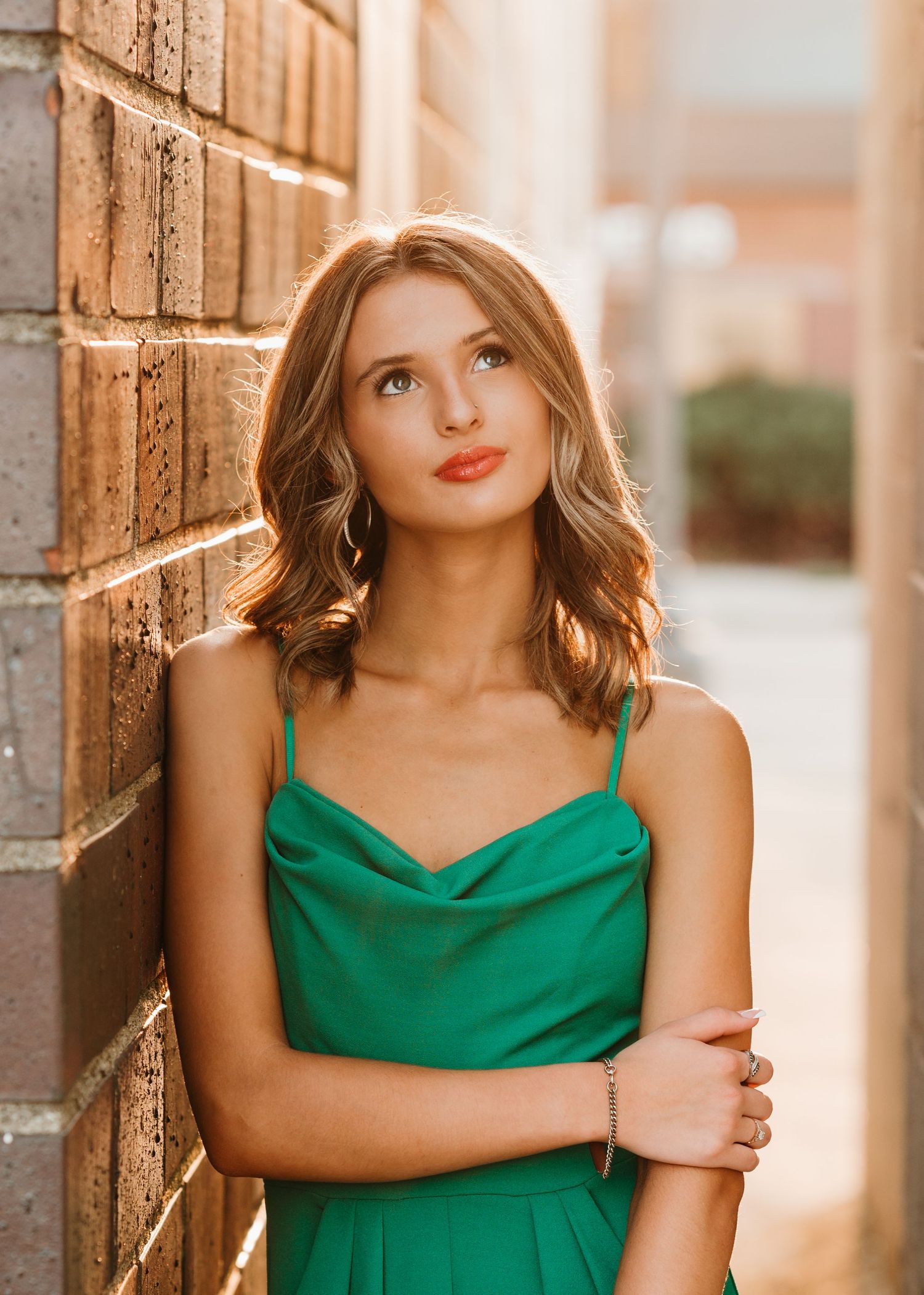 Portrait in emerald green dress against brick wall with warm sunset lighting.