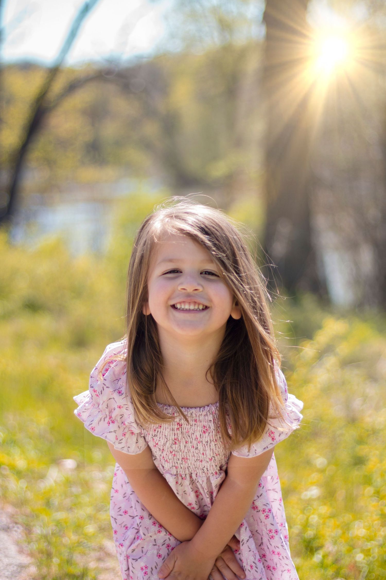 Child smiling joyfully while standing in a sunlit field near a gently flowing river, surrounded by tall grass and a peac