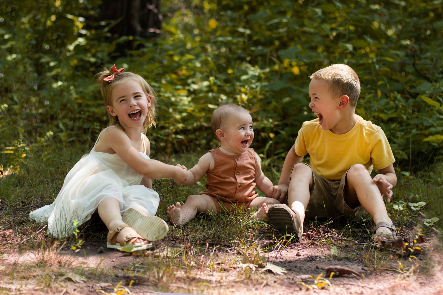 Three young siblings laughing hysterically together, their faces filled with pure joy and uncontainable giggles, capturi