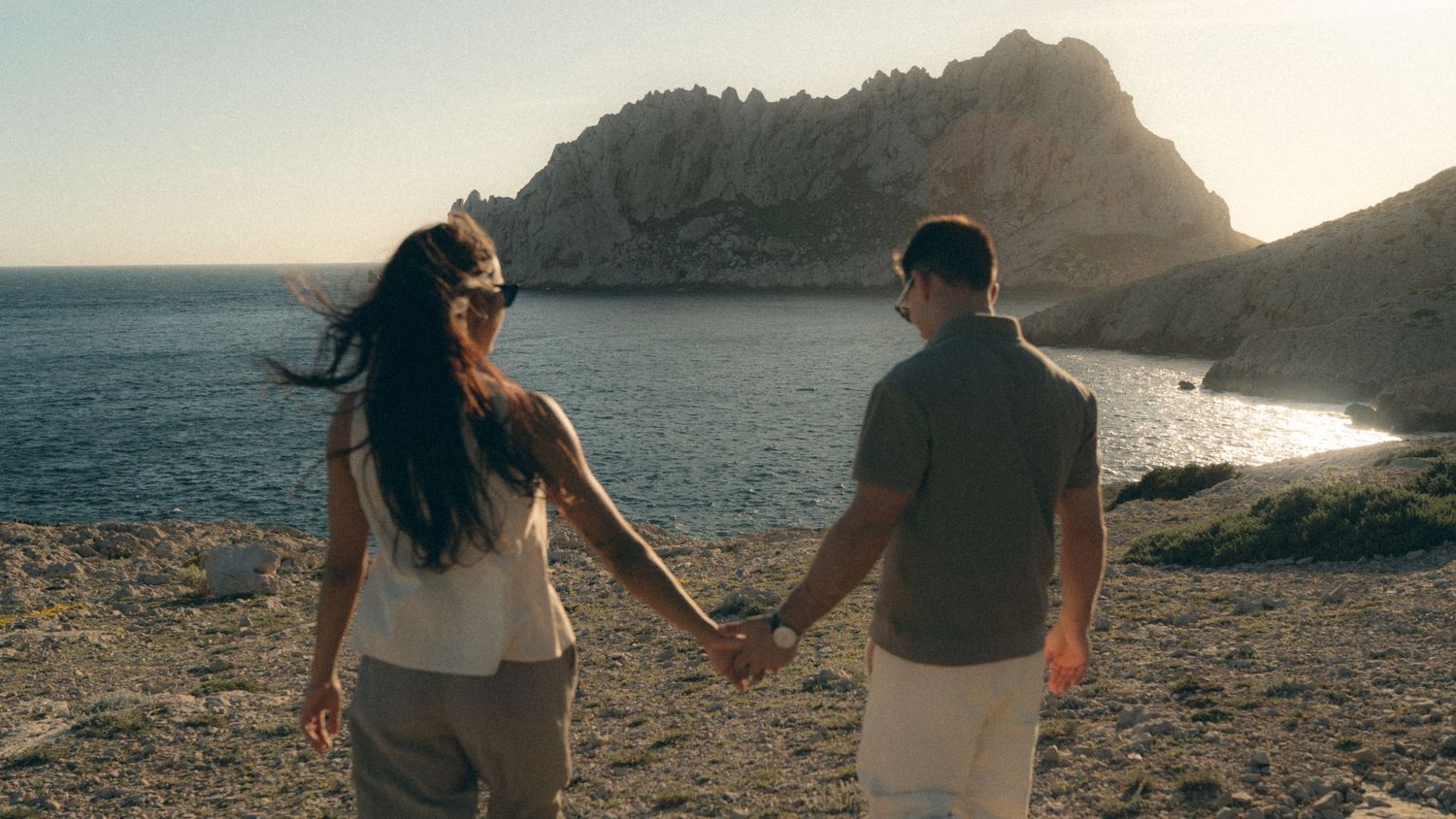 Couple holding hands while walking along coastal cliff path at sunset.