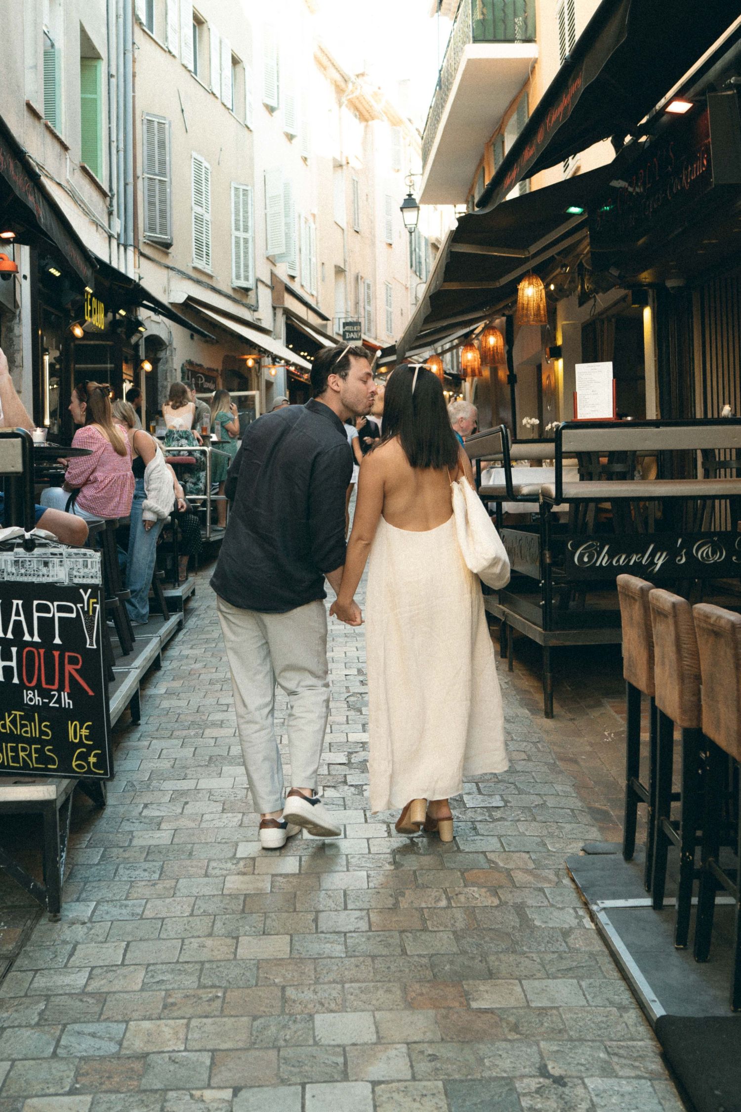A couple strolls down a charming cobblestone street lined with cafes and shops in a European city.