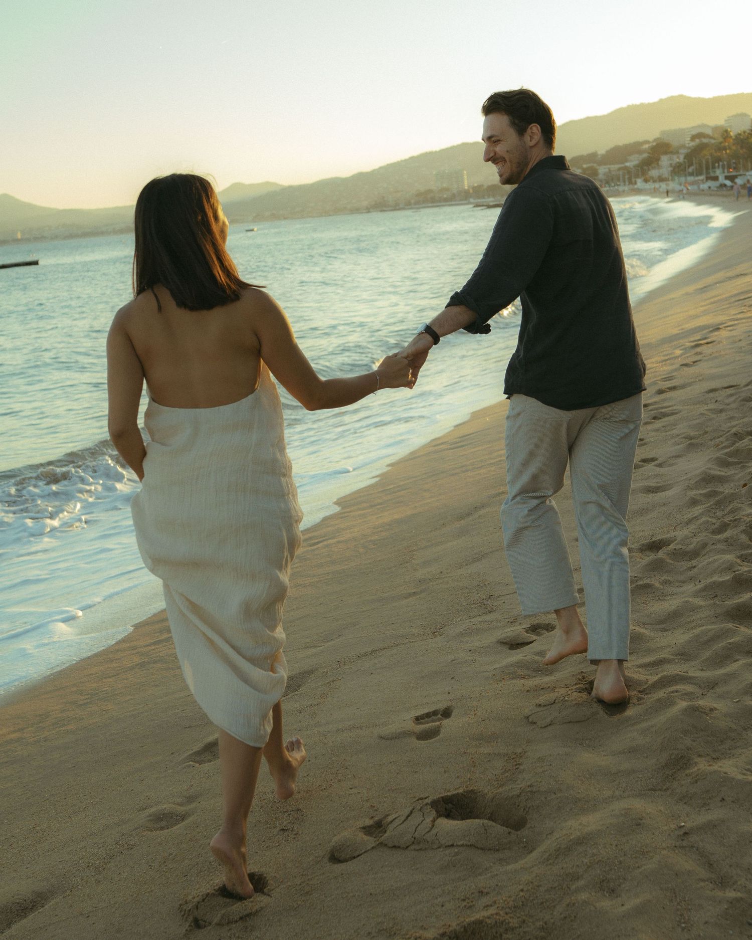 A couple in casual attire walks playfully along a scenic beach shoreline at sunset.