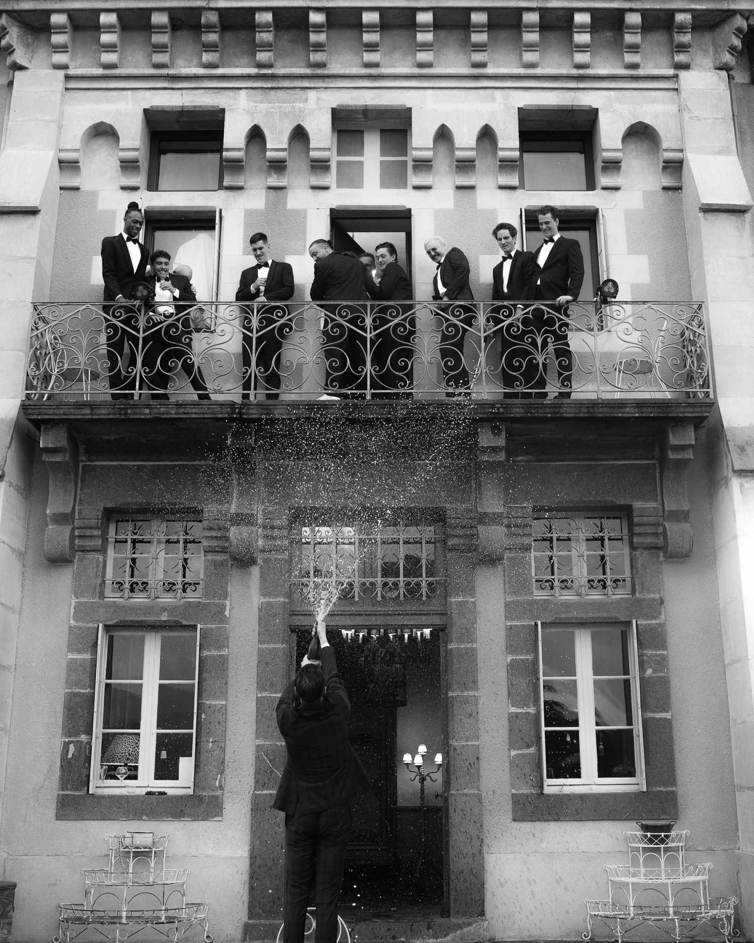 A group of formally dressed people stand on a balcony of a historic stone building with ornate railings.