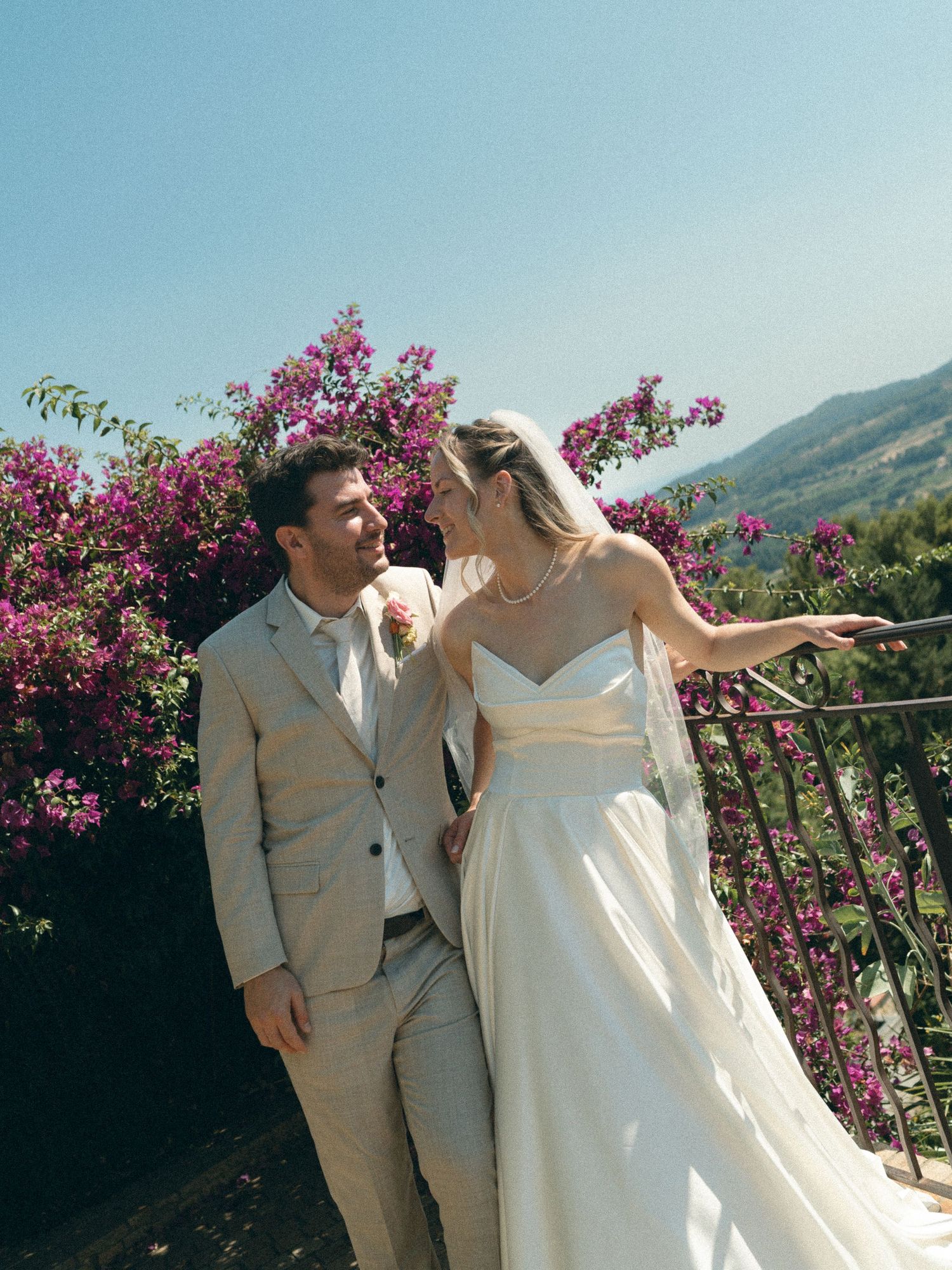 Bride and groom share a romantic moment on a balcony surrounded by pink bougainvillea with mountain views.