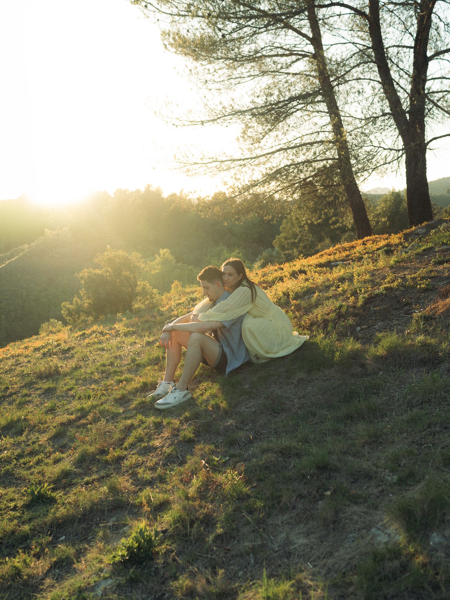 Two people embracing on a sunlit grassy hillside at golden hour, with trees in the background.