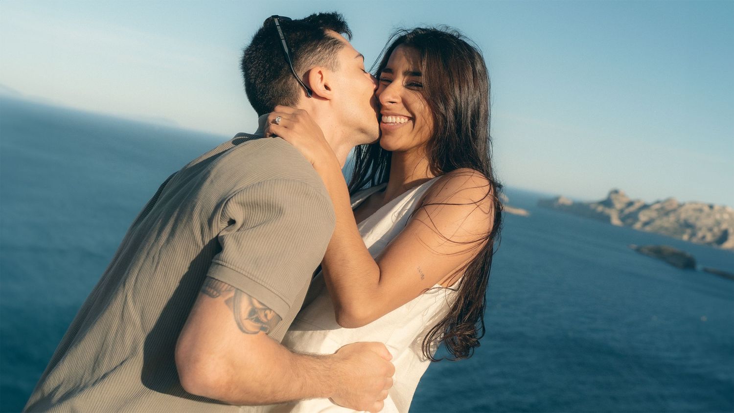 A romantic couple embraces on a cliff overlooking the ocean during sunset creating a silhouetted moment.