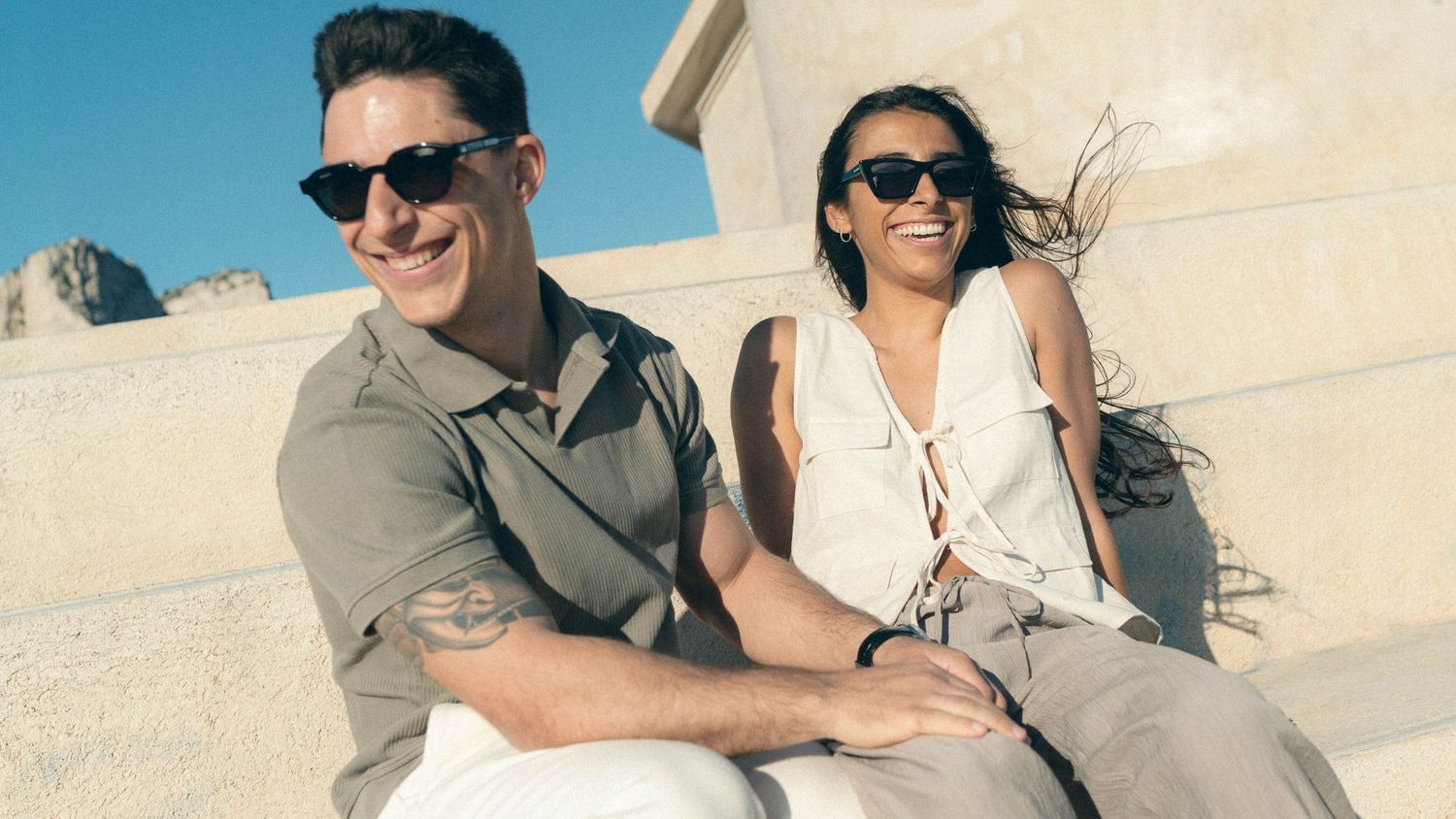 Two people in sunglasses sit together smiling outdoors on a sunny day wearing casual summer attire.